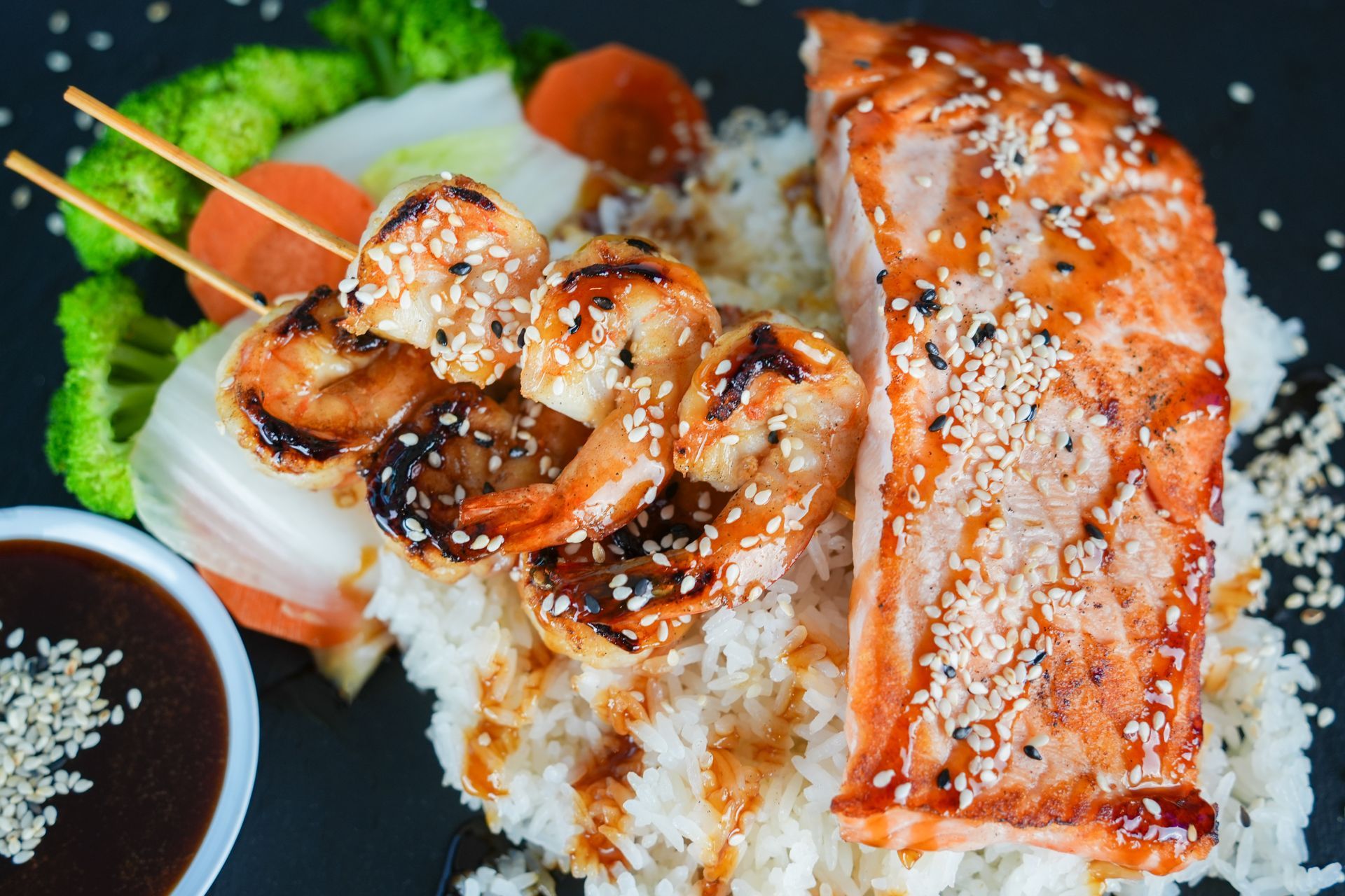 Grilled shrimp and salmon over rice with dipping sauce on a black plate