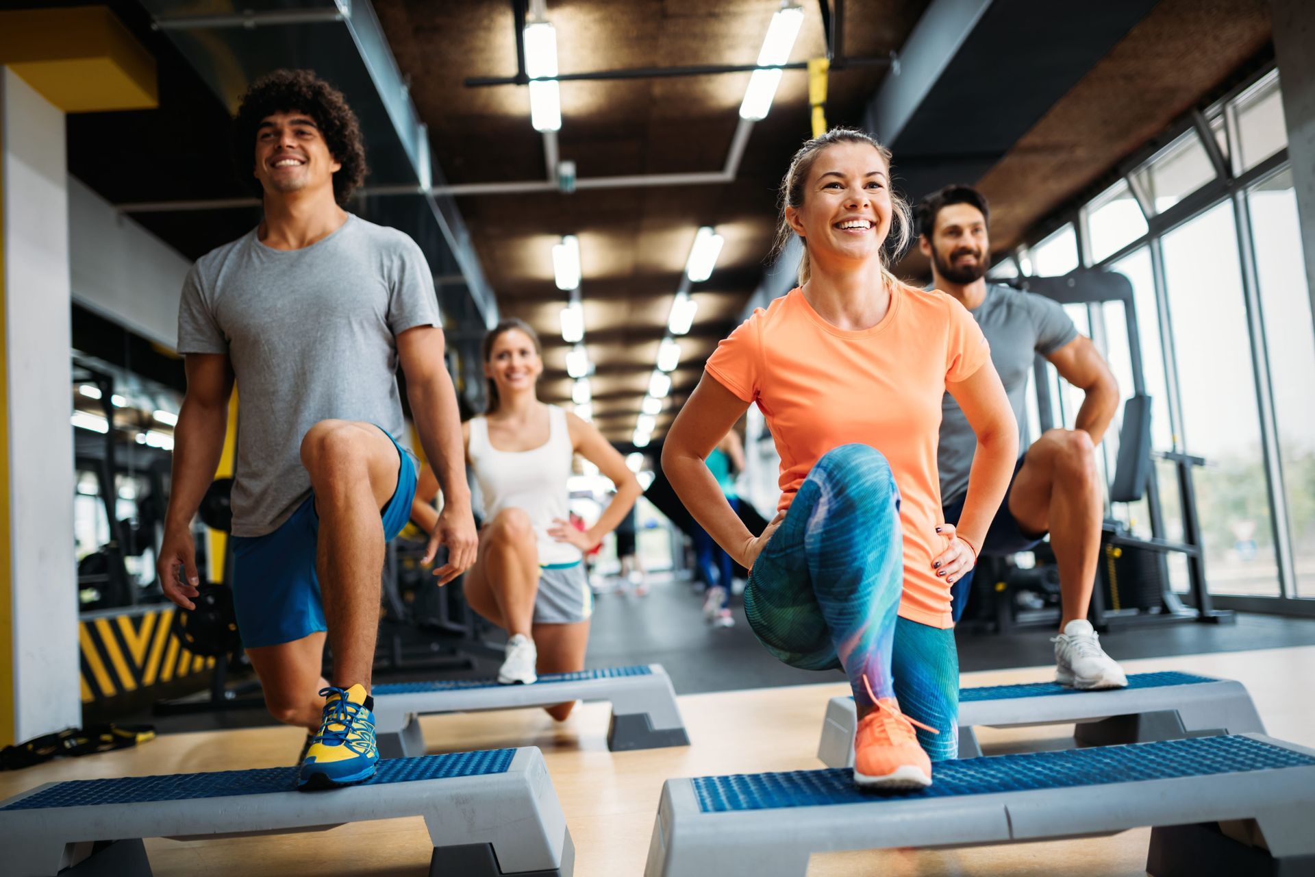 Four people exercising on steps in a gym. They are smiling and lunging.