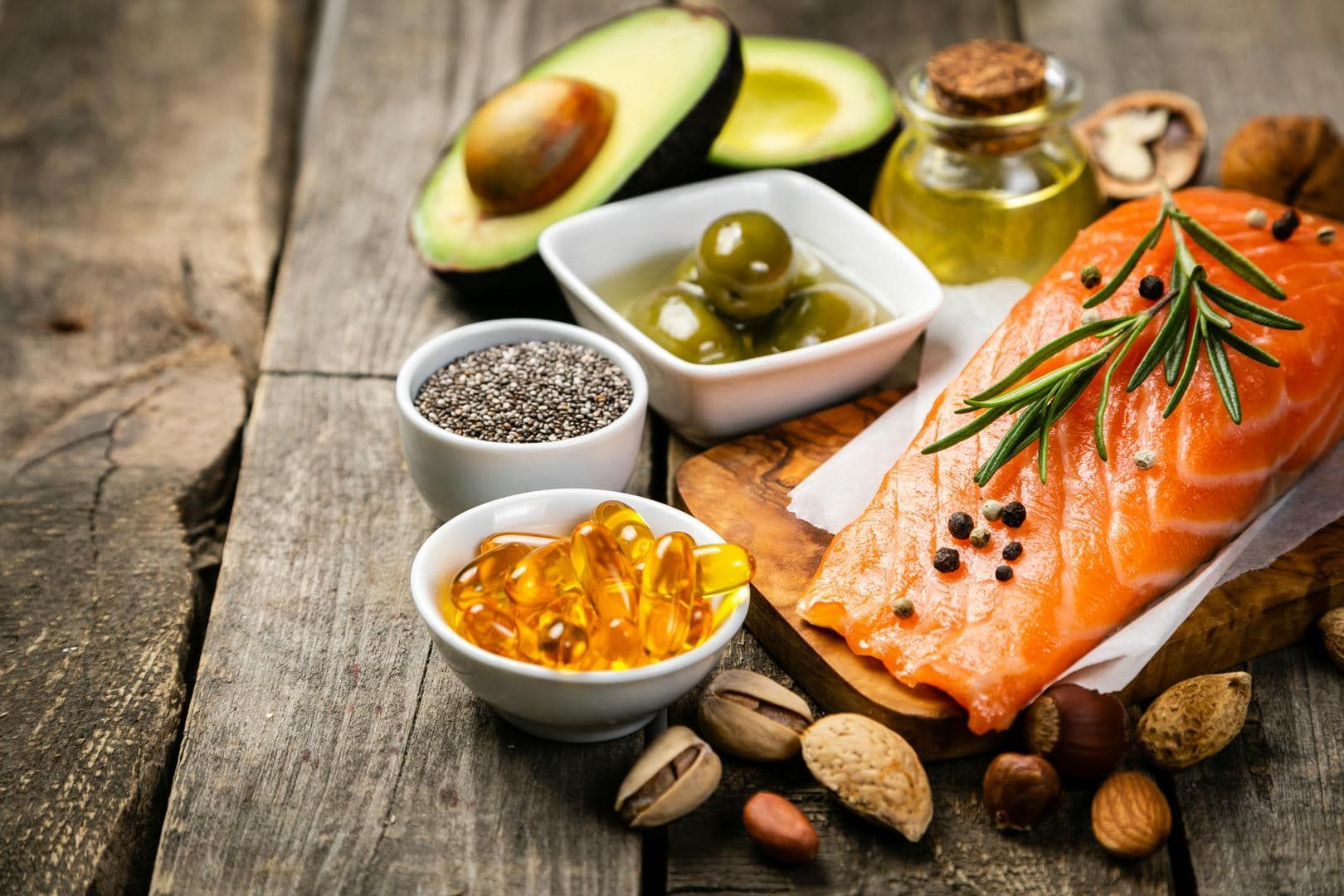 Assortment of healthy fats: avocado, salmon, nuts, olive oil, chia seeds, and olives on a wooden surface.