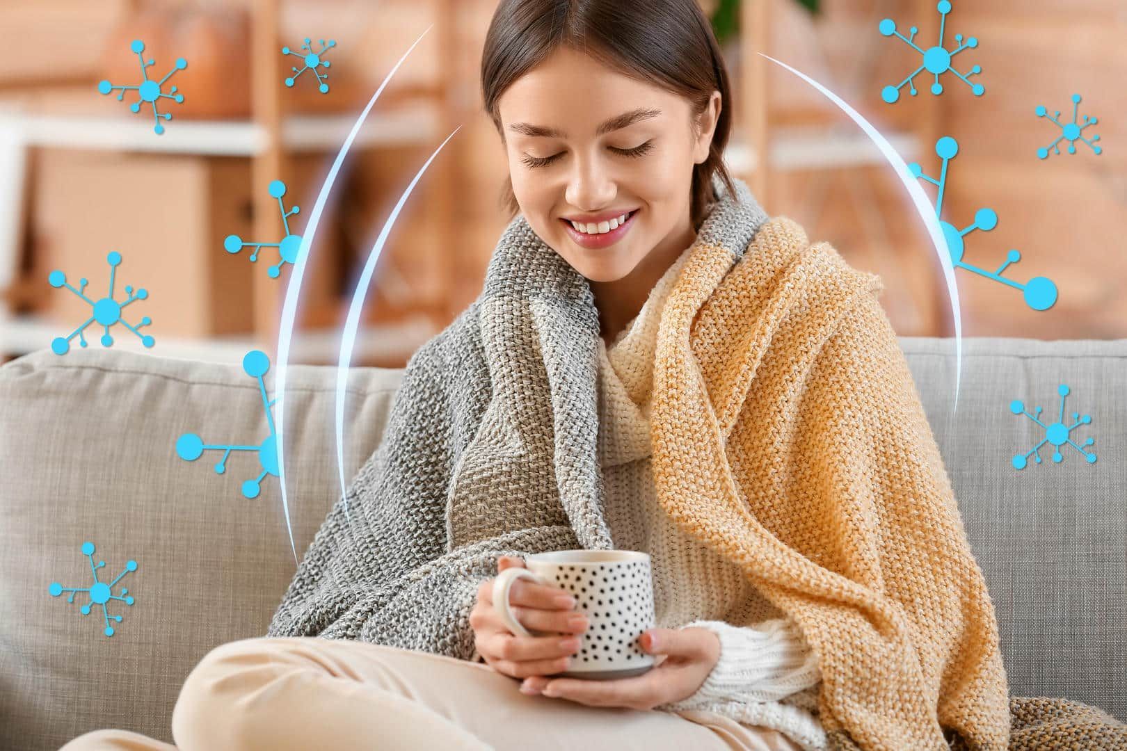 Woman wrapped in blanket, holding a mug, smiling, surrounded by illustrated blue virus particles.
