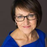 A woman wearing glasses and a blue shirt is smiling for the camera.