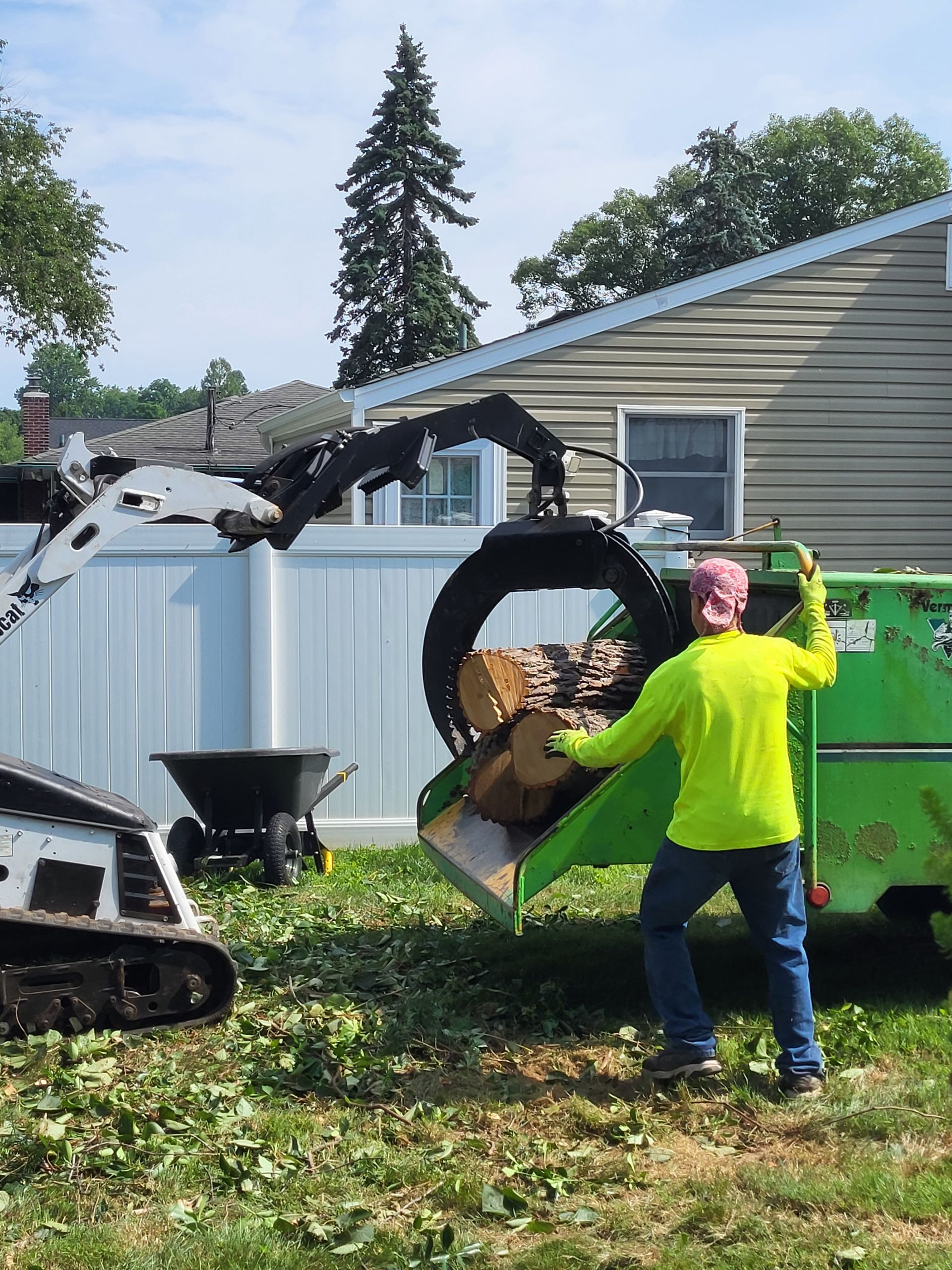Person in neon green jacket using excavator to lift debris beside a house and white fence