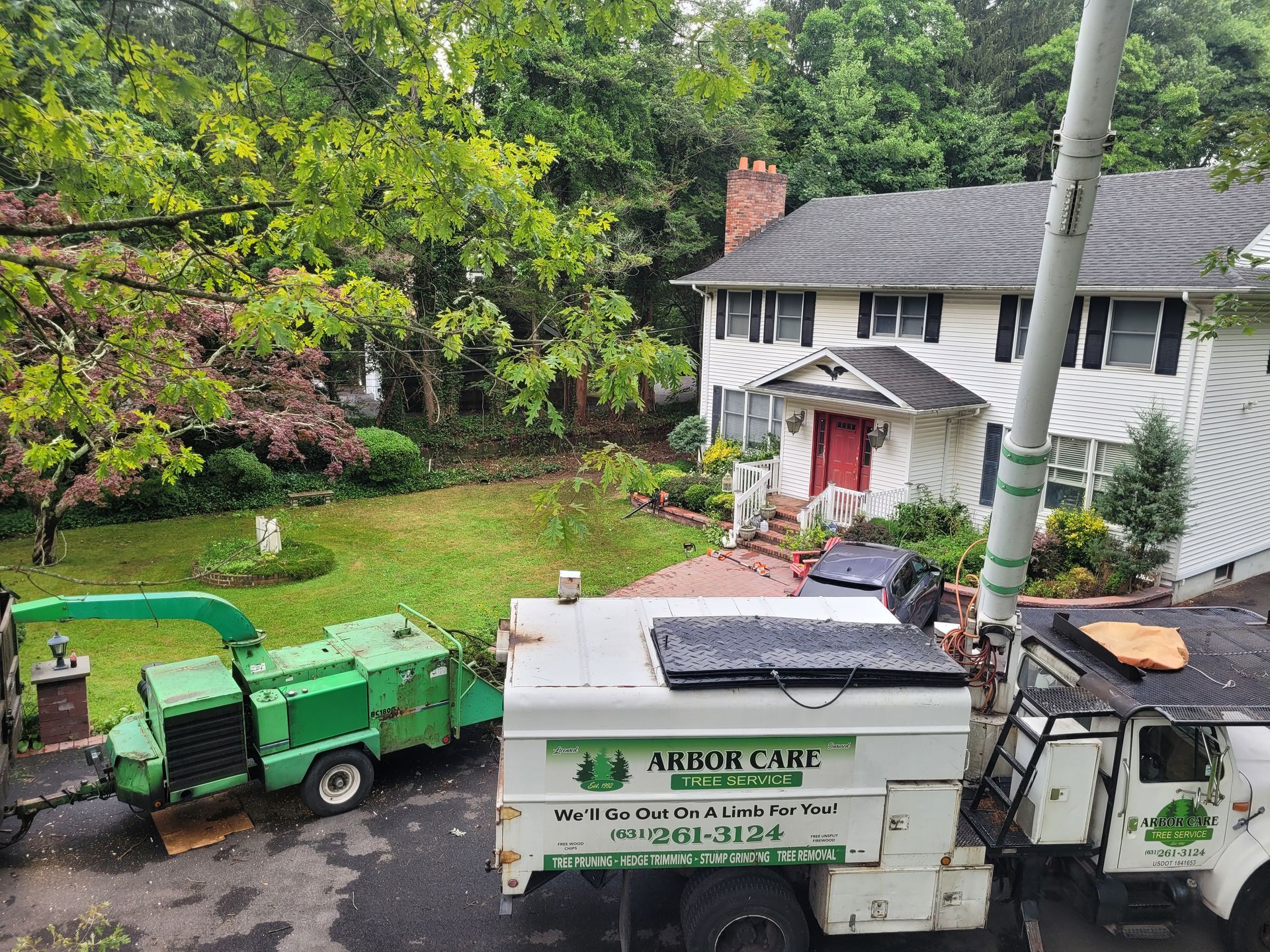Two trucks and a cherry picker outside a white house with a red door, surrounded by trees and yard debris