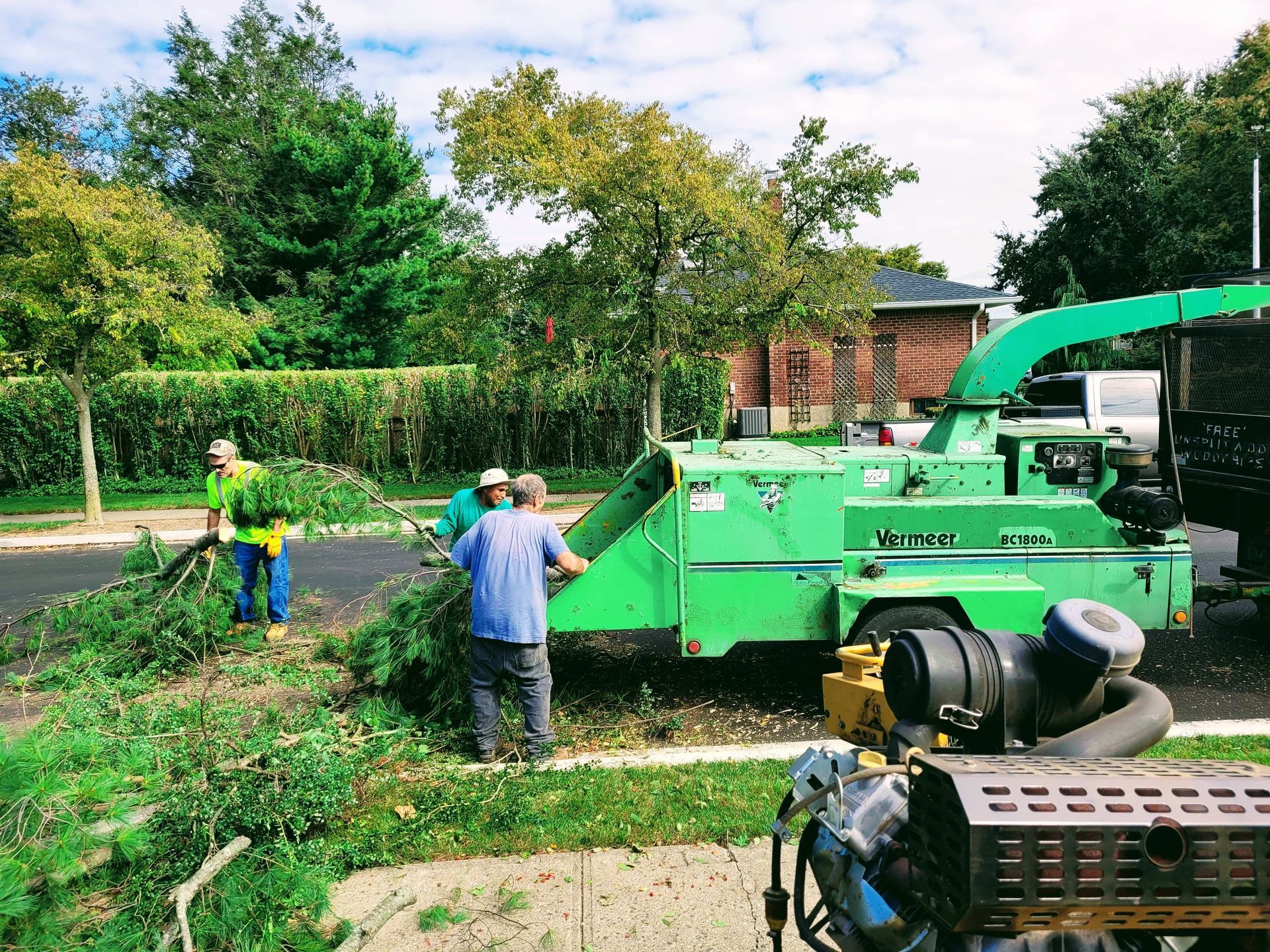 Workers clearing leaves beside a green industrial machine in a sunny park-like setting