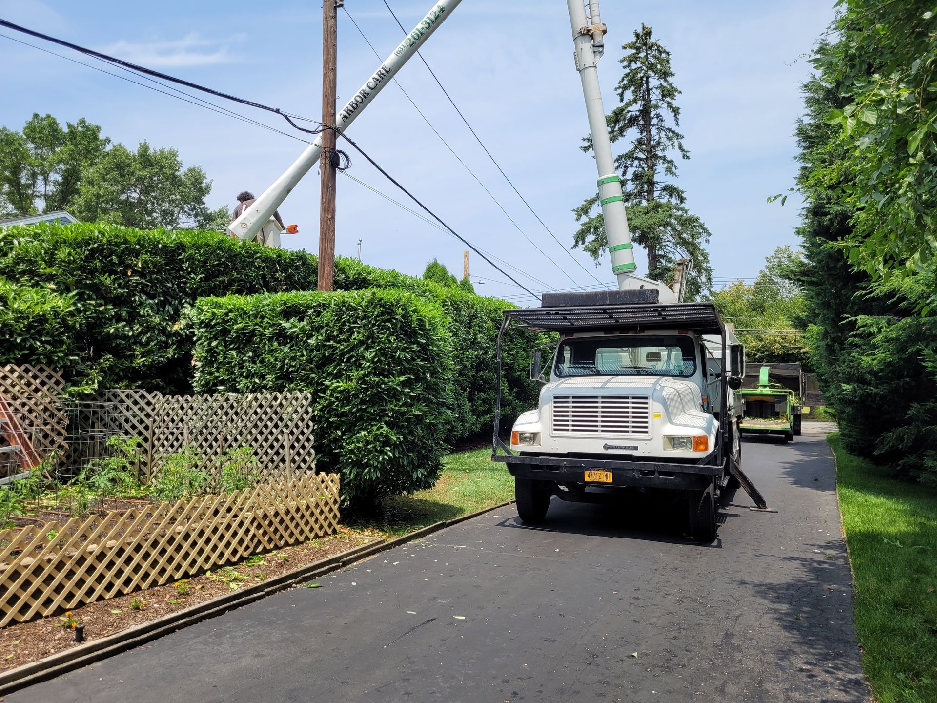 Utility truck on a narrow driveway beside hedges and power lines, with another truck in the background.