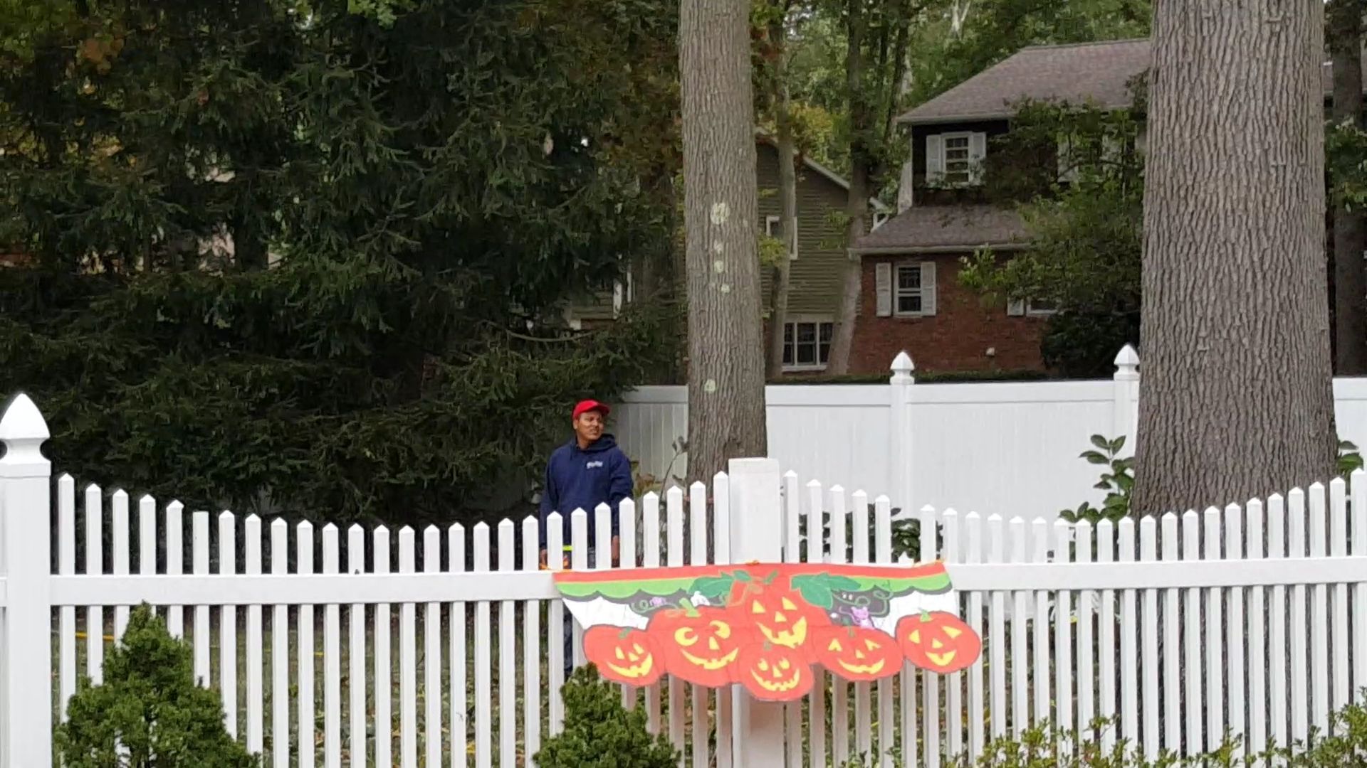 Person behind a white picket fence near a house, with orange pumpkin decorations on the fence.
