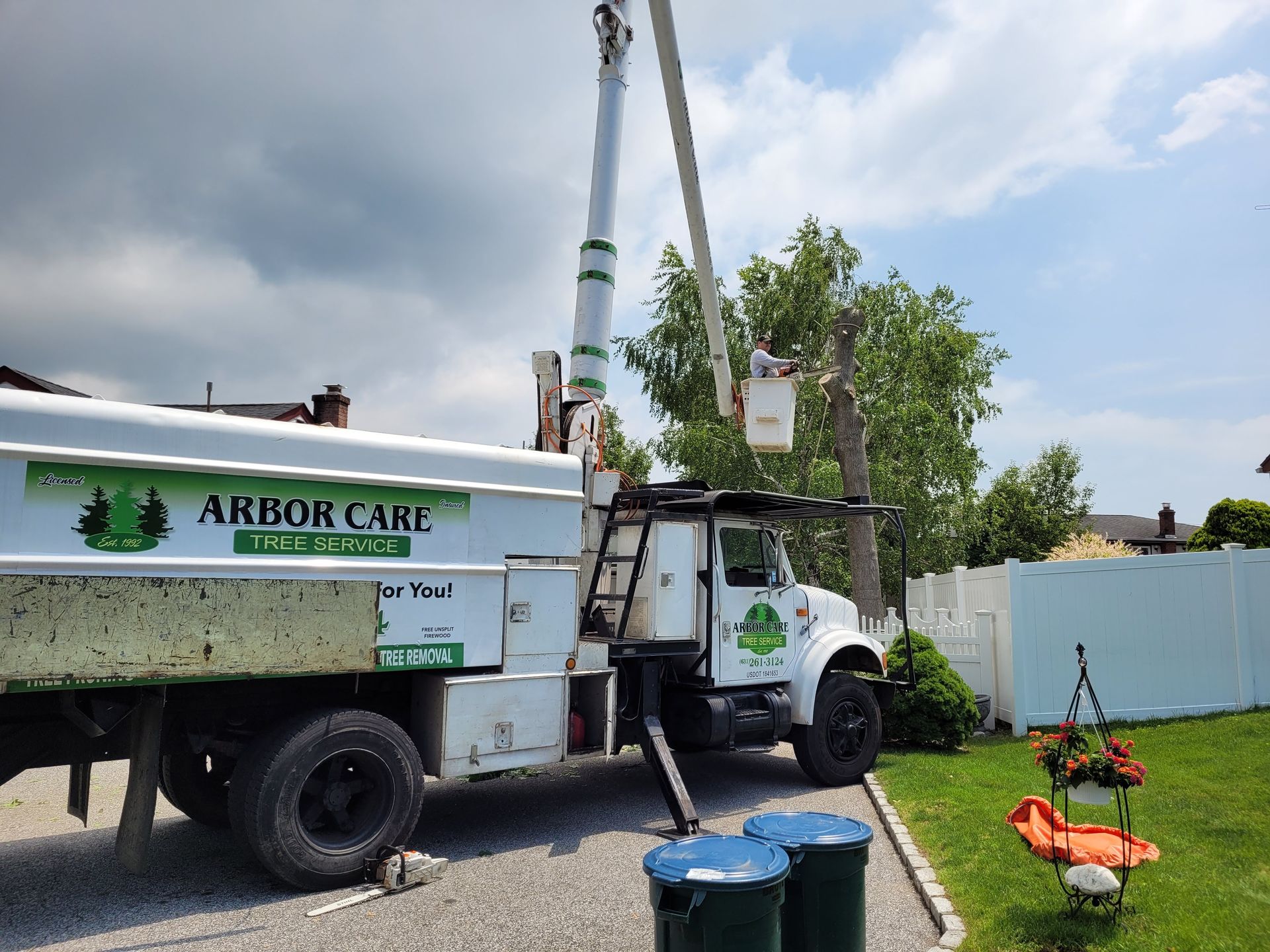 Arbor Care tree service truck with crane lifting equipment beside a fenced yard and orange chipper trailer