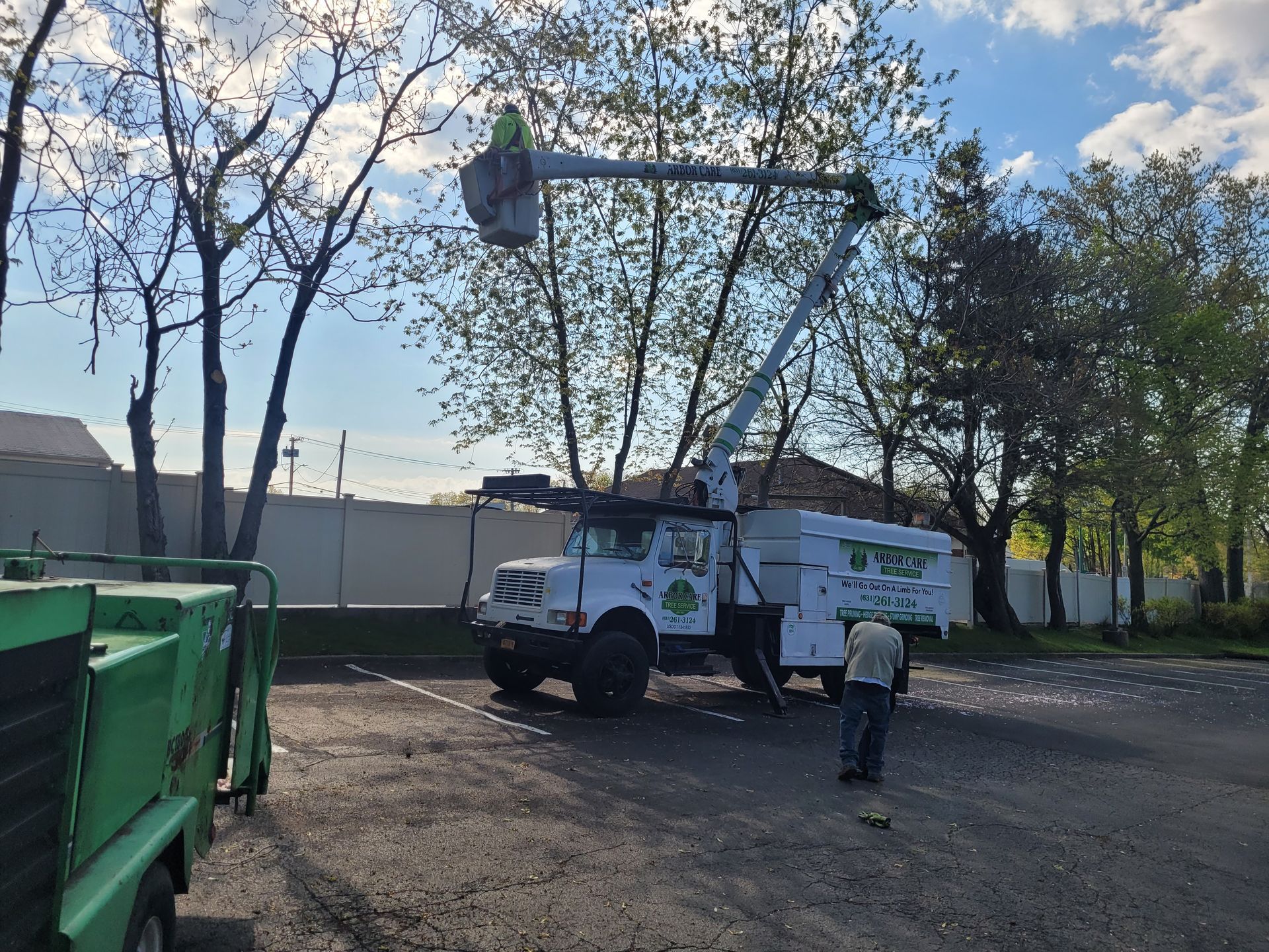 Utility truck and worker using a bucket lift beside a green dumpster on a tree-lined street.