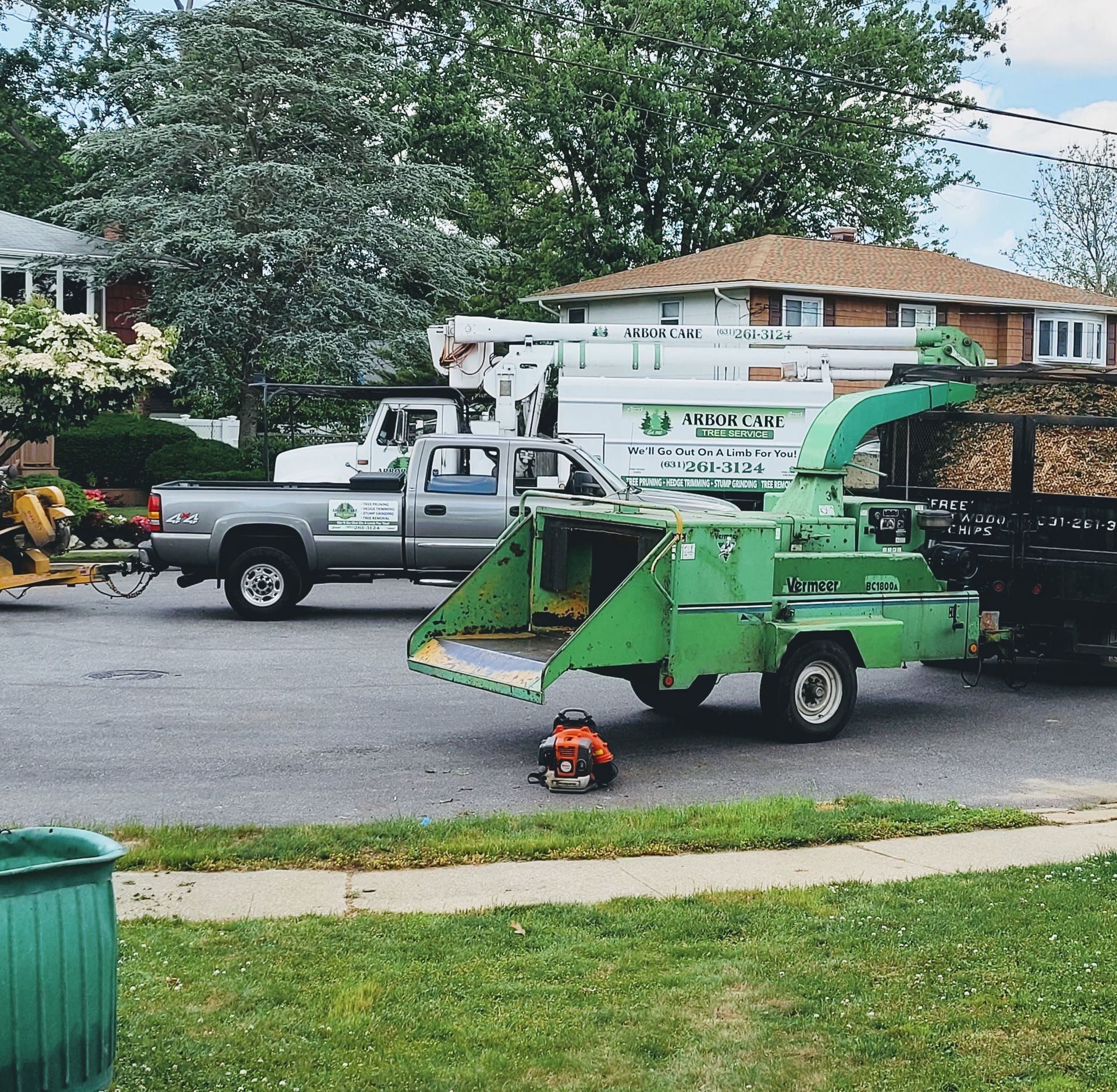 Green wood chipper parked on a residential street beside a truck, with a worker nearby and cut logs loaded behind.