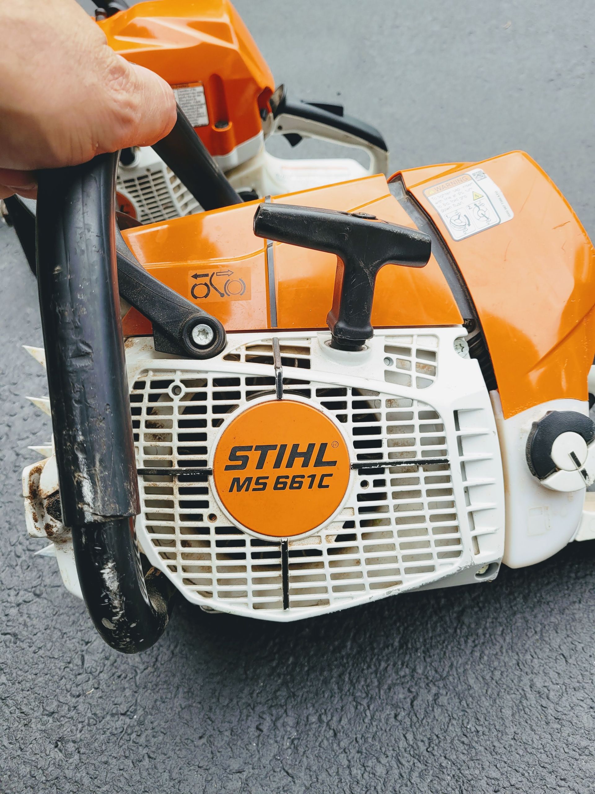 Close-up of an orange STIHL chainsaw being held by a hand on a white surface.