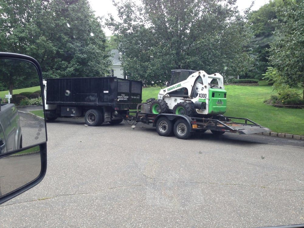 White skid steer loaded on a trailer beside a black dump trailer in a tree-lined parking lot