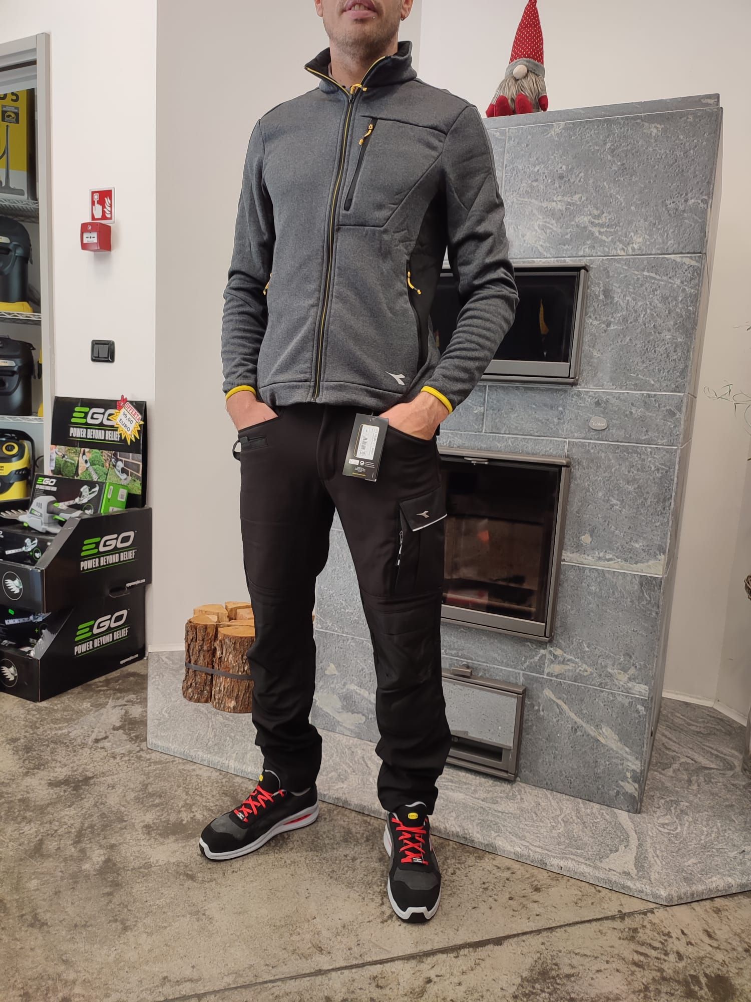 Un uomo è in piedi in un ambiente interno, accanto a un camino rivestito di piastrelle grigie, e indossa una giacca grigia, pantaloni neri e scarpe da ginnastica rosse e nere.