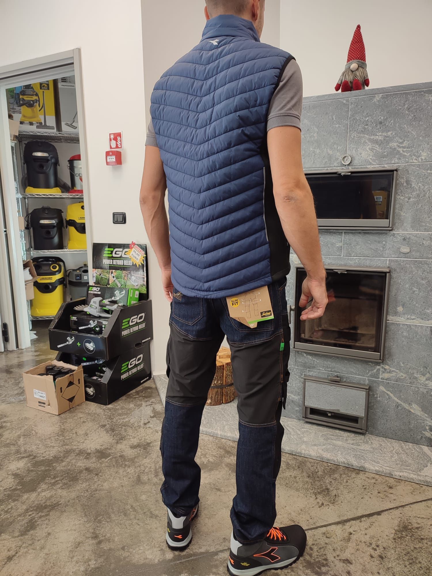 Persona in piedi in un ambiente interno, vicino a un camino in cemento, con indosso un gilet blu, una camicia grigia, jeans e scarpe da ginnastica nere.