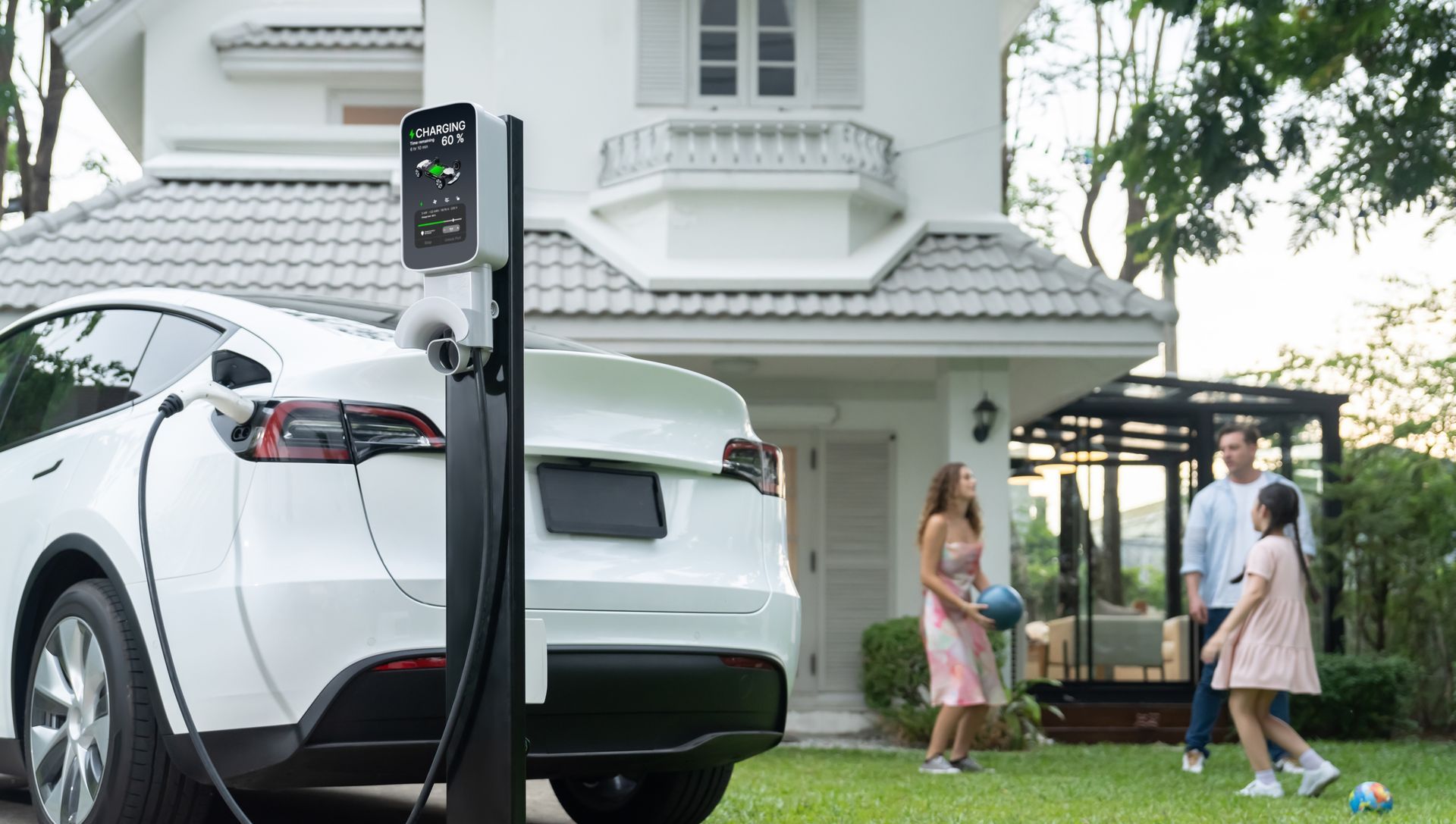 White electric SUV charging beside a house while two people stand nearby on the lawn
