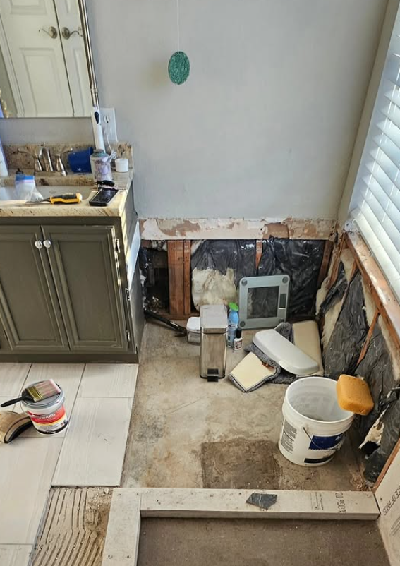 Kitchen wall opened up to studs during renovation, with debris, tools, and a bucket on the floor.
