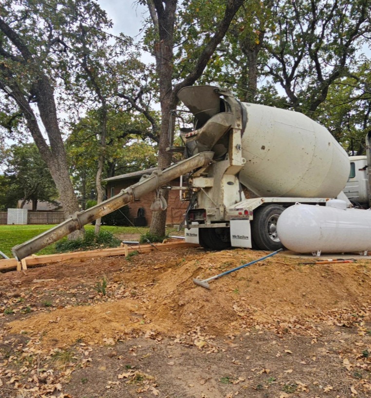 Concrete mixer truck pouring concrete into a dirt area on a wooded residential lot.