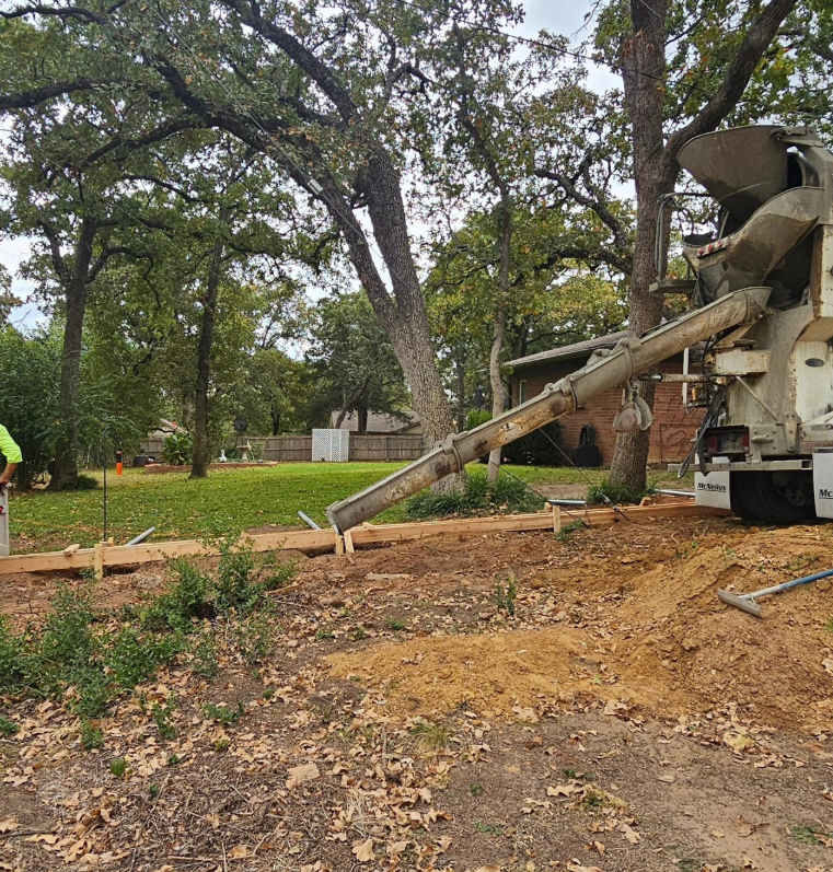 Crew pours concrete in a wooded yard beside a cement truck and chute.