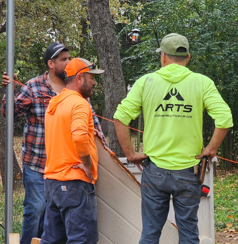 Three workers in neon safety gear and a plaid shirt lifting a panel outdoors near trees and a truck