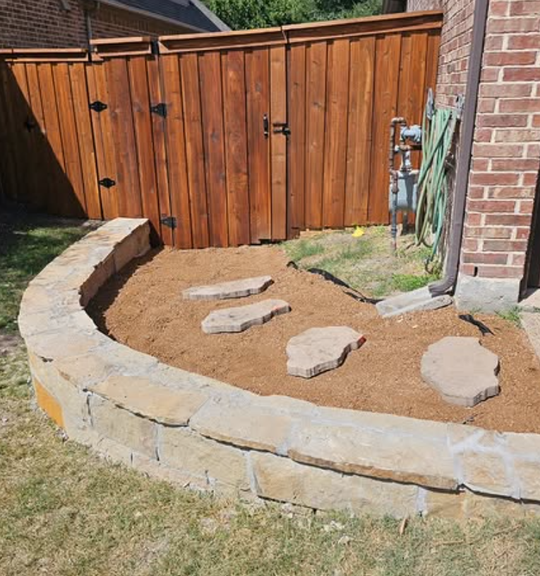 Curved stone garden bed with mulch and stepping stones beside a wooden fence and brick house.