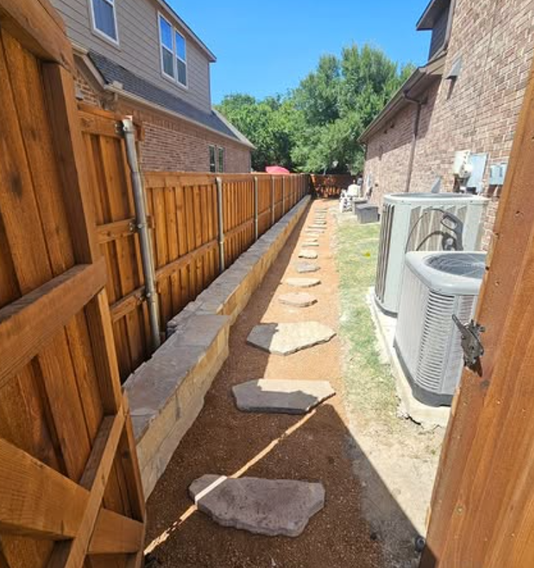 Narrow gravel pathway between brick houses with a wooden fence and AC units, lined with stepping stones.