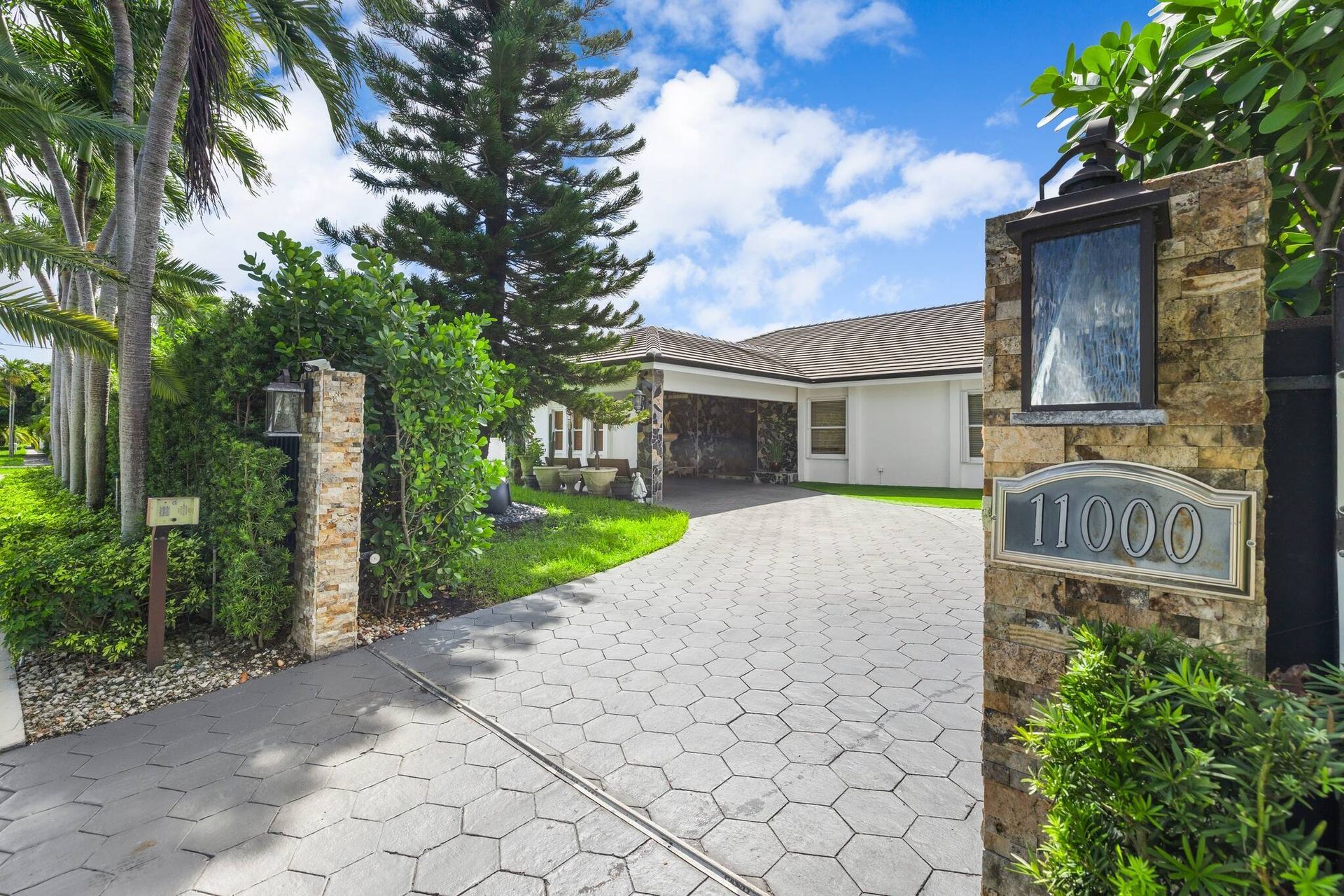Entrada privada con portón que conduce a una casa, con palmeras, pilares de piedra y un cielo azul intenso