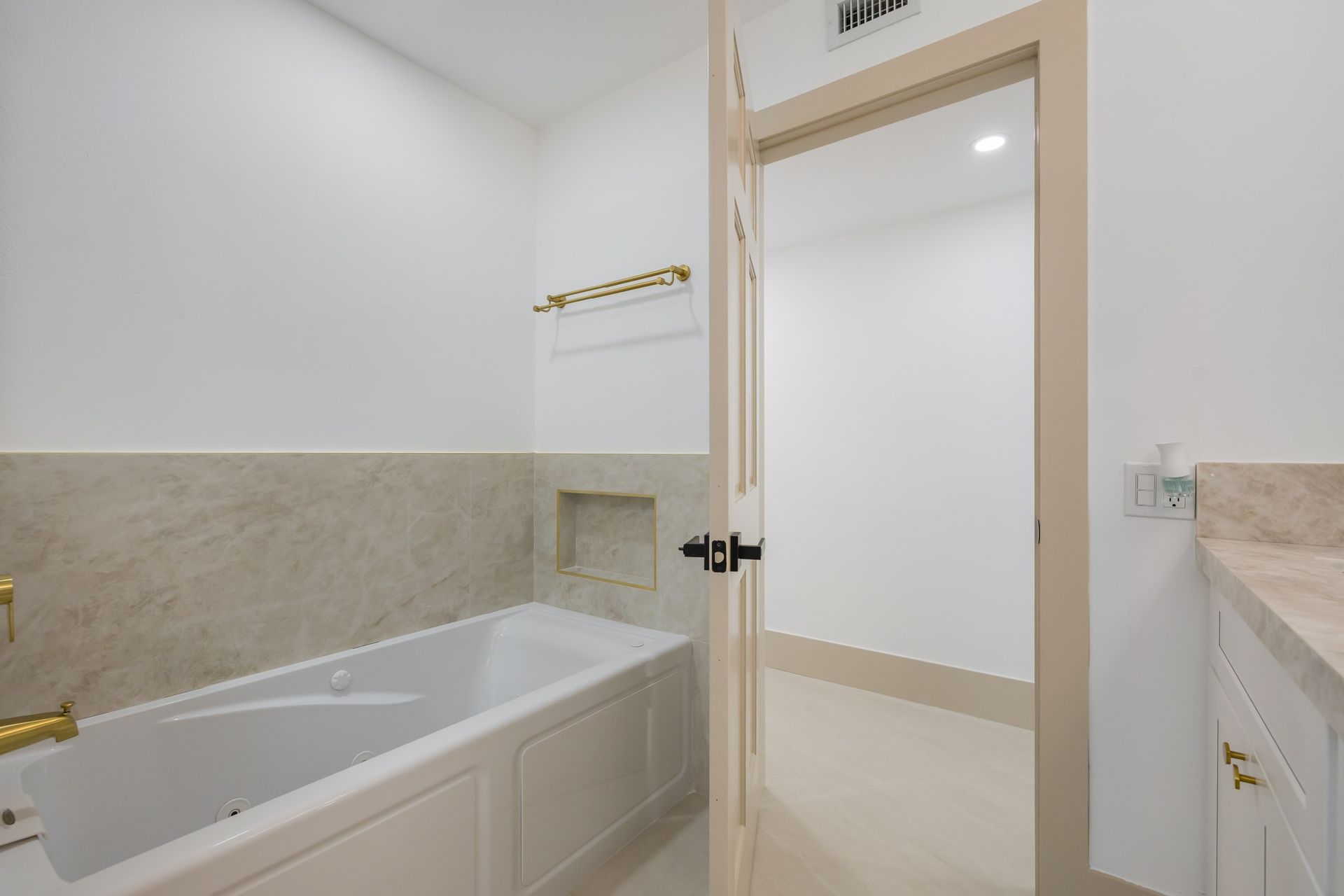 Bright bathroom with a bathtub, beige tile, and an open door to a white adjoining room.