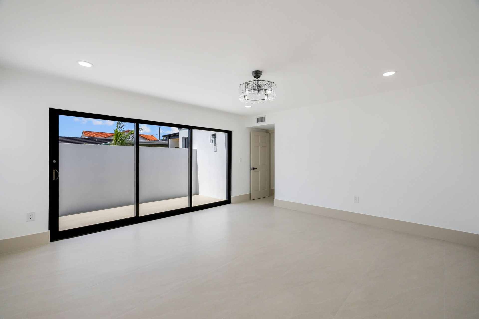 Bright empty living room with white walls, beige floors, and large black-framed sliding glass doors.