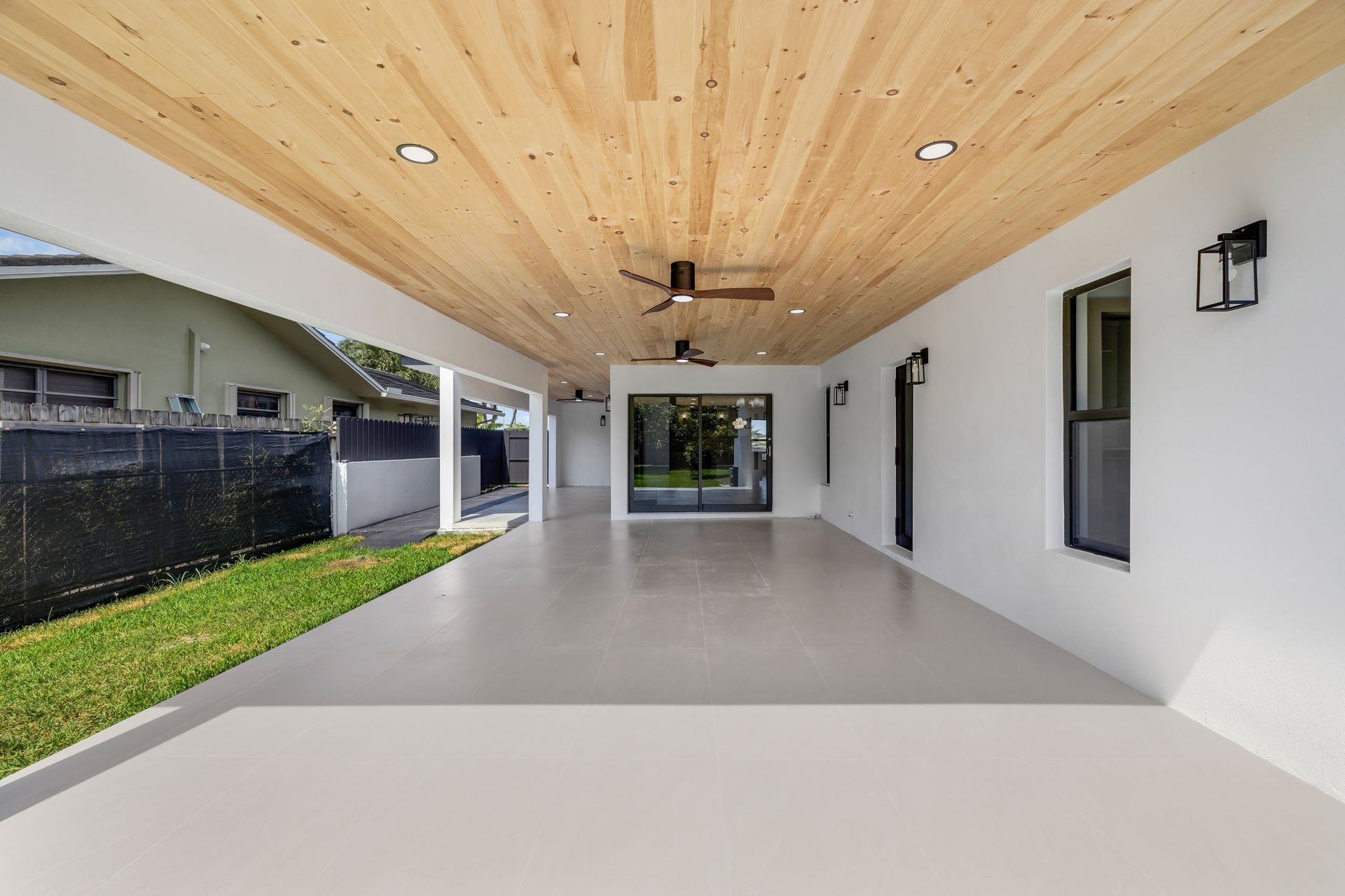 Modern covered patio with white walls, wood ceiling, sliding doors, and a small lawn along the side