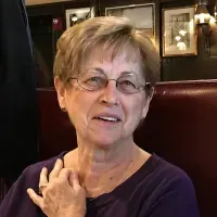 Smiling woman with short hair and glasses, wearing a purple top, seated in a restaurant booth