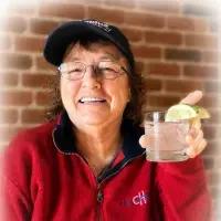 Smiling person in a red jacket and black cap holding a drink with a lime wedge