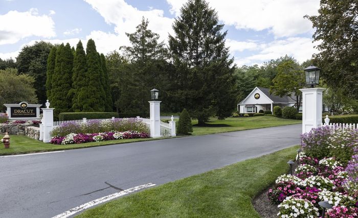 Driveway entrance with white pillars, pink flower beds, and a house in the distance under cloudy skies