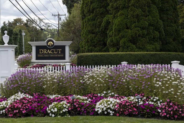 Tracuit entrance sign with white fence and purple-and-white flowers in front of tall evergreens