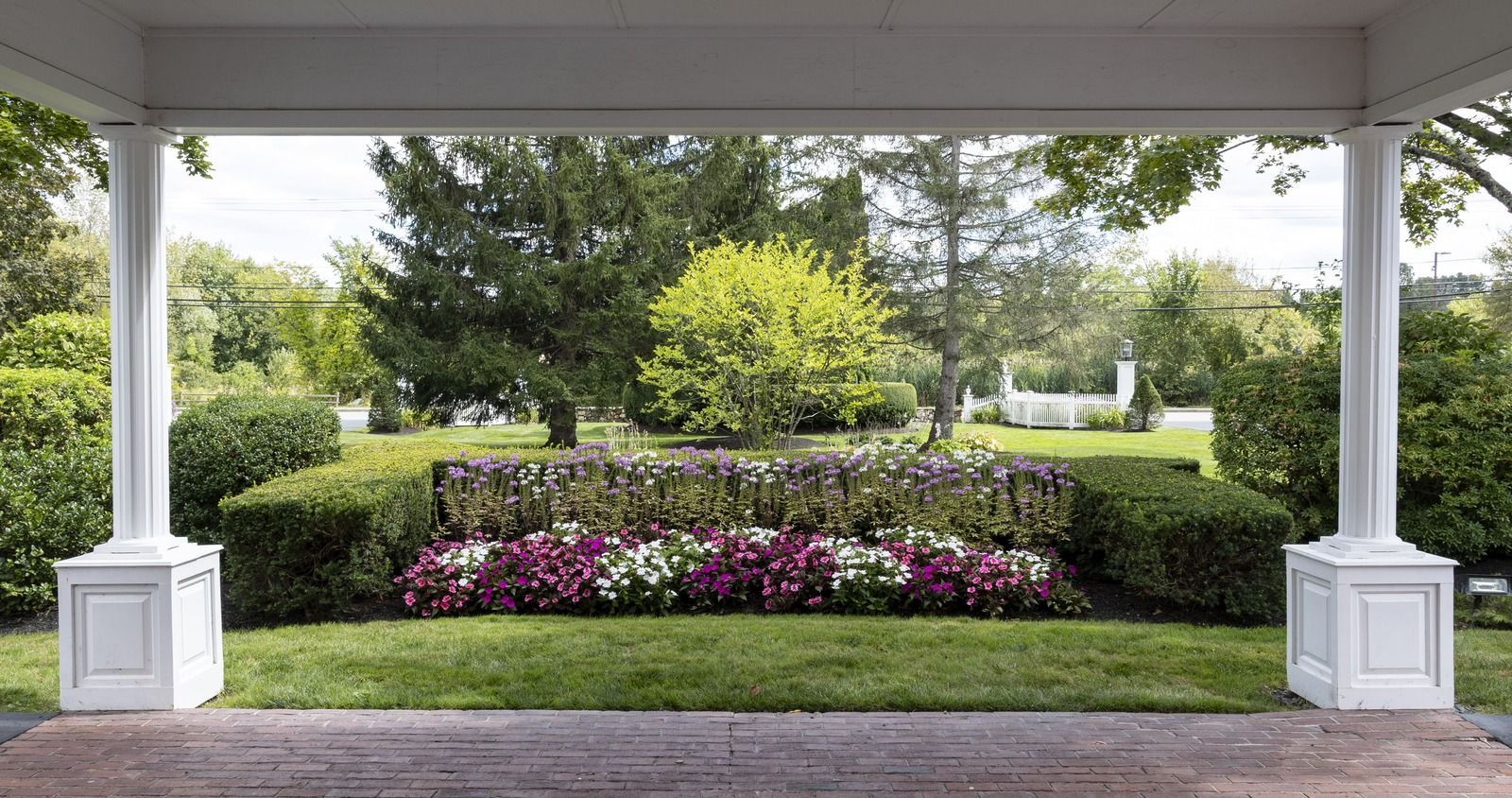 Covered porch framing a landscaped garden with white columns, purple flowers, hedges, and trees beyond