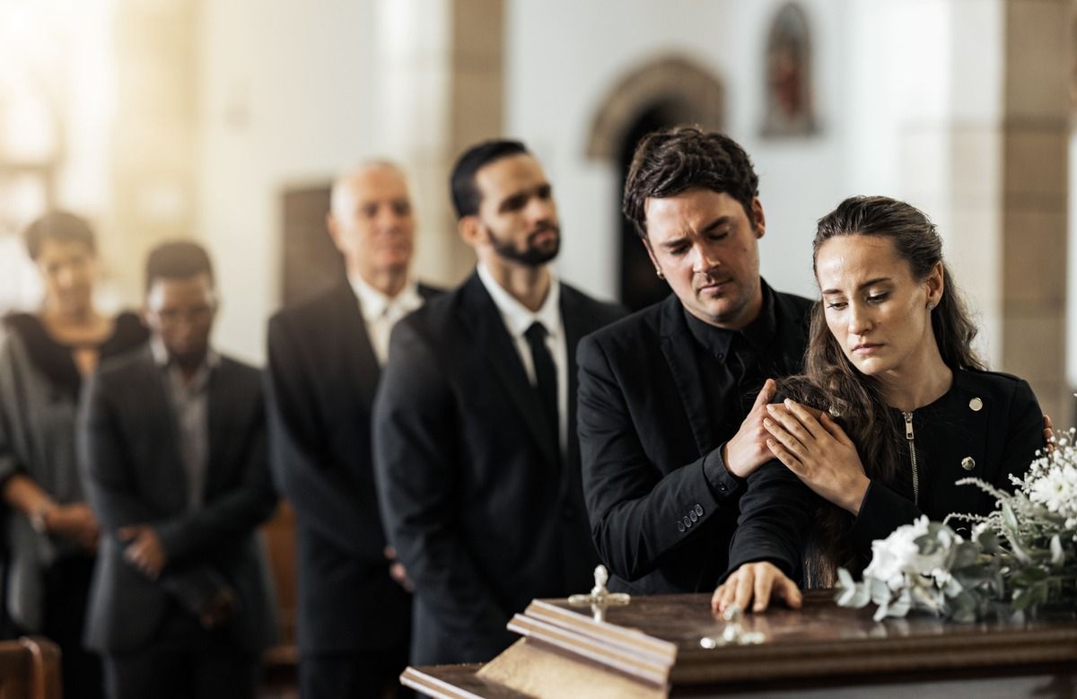 Mourners stand beside an open casket at a funeral, with a grieving woman leaning on a man’s shoulder.