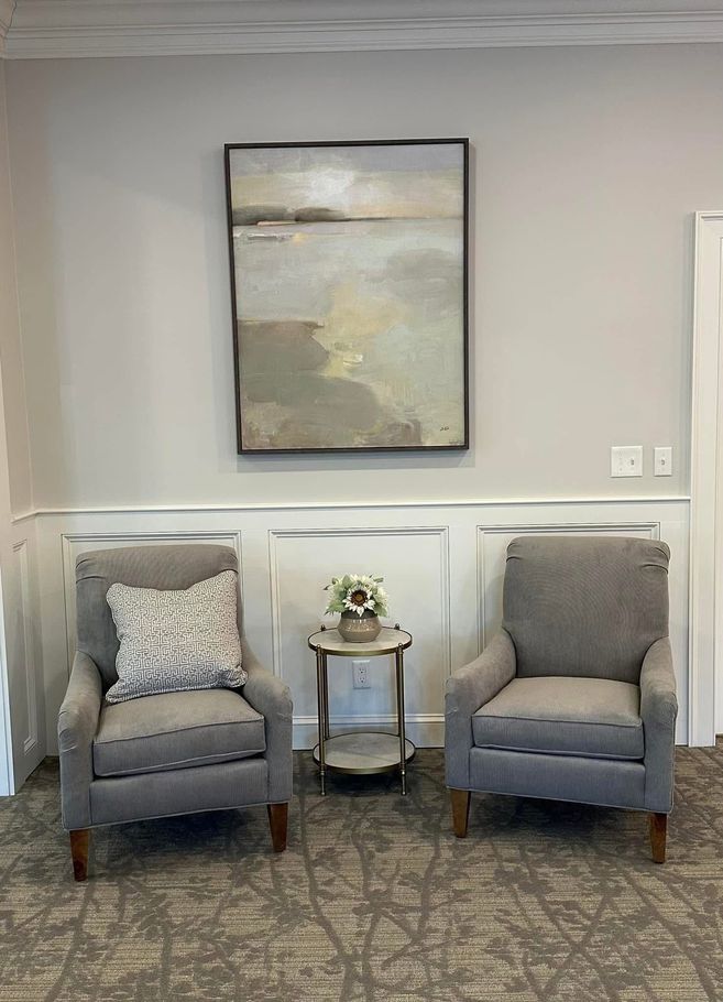 Two gray armchairs with a small side table beneath framed wall art in a neutral lounge.