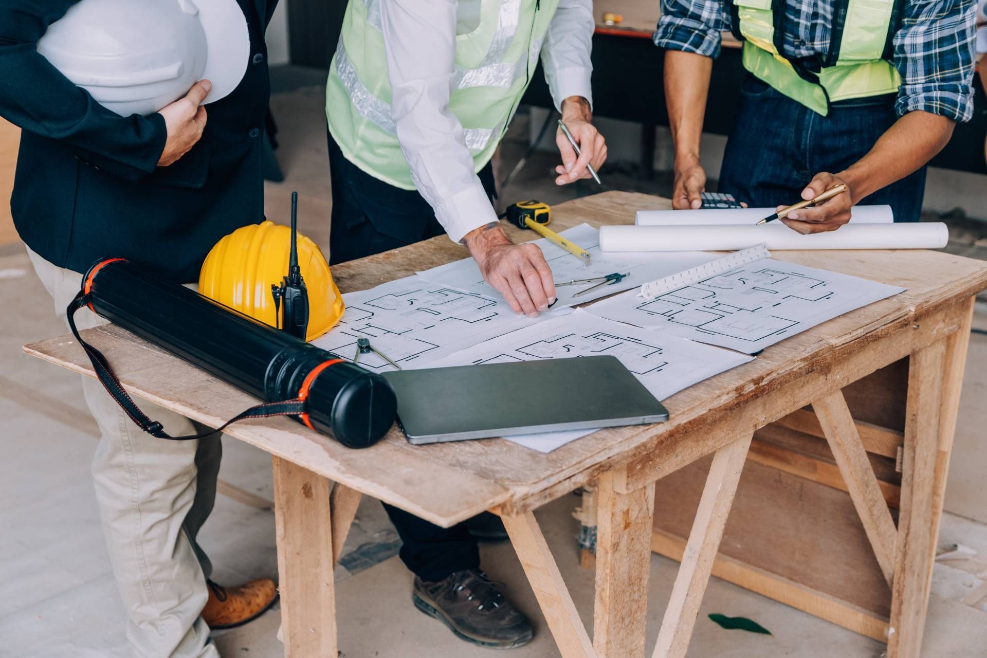 People reviewing architectural plans at a wooden table, with a hard hat and rolled blueprints nearby.