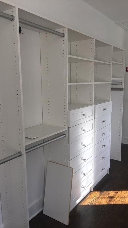 White built-in closet with shelves, drawers, and hanging rods, one cabinet door open, in a sunlit room