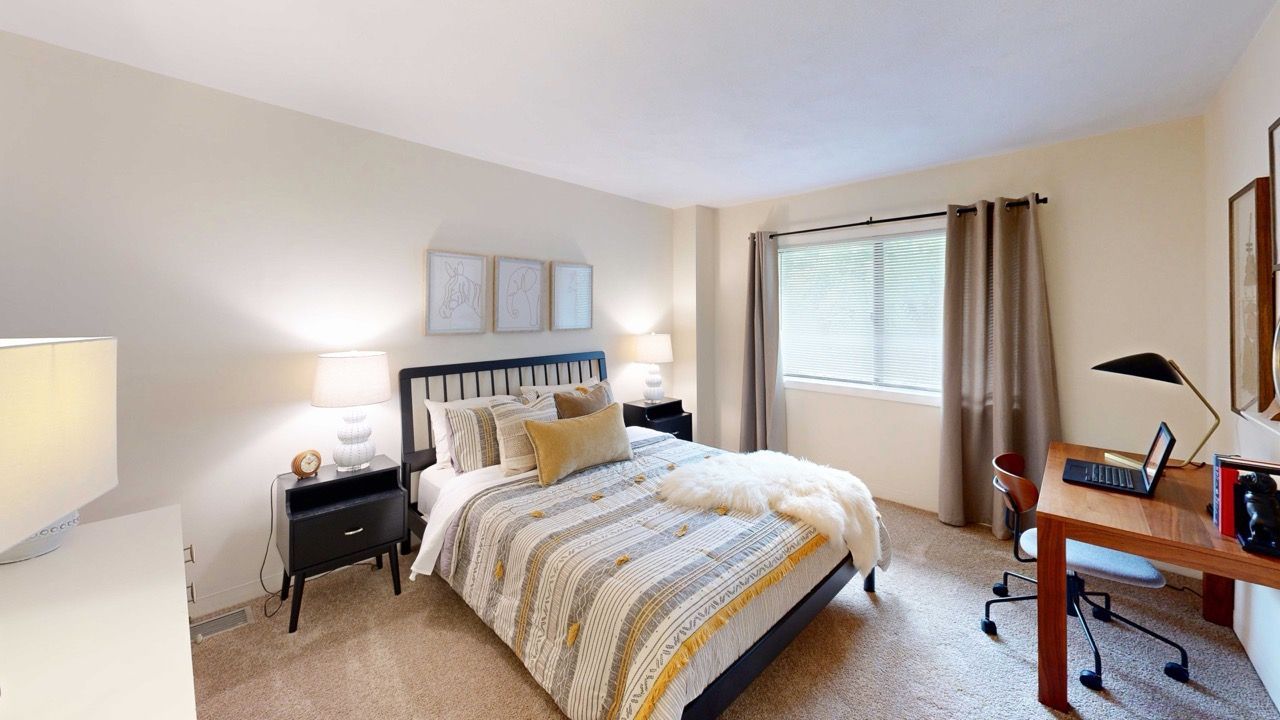 Bedroom in an apartment with a queen bed, two nightstands, a wooden desk, and a large window with curtains.