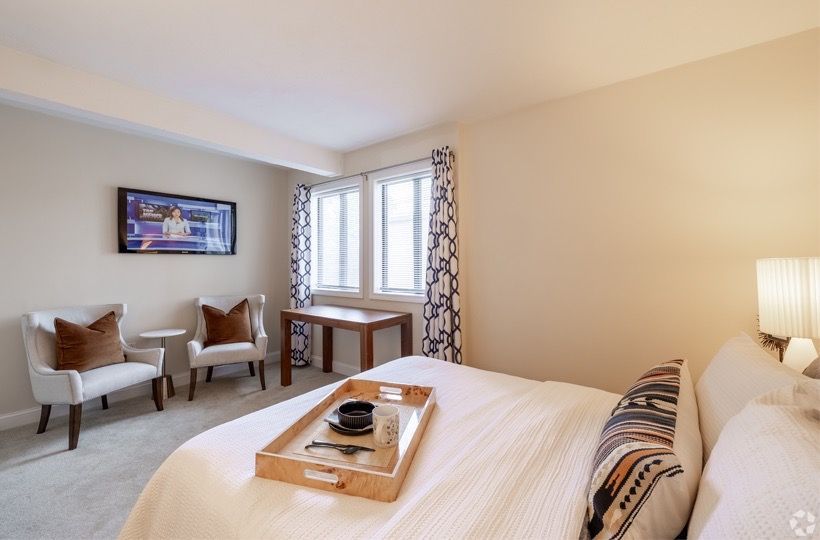 Hotel-style bedroom with a large bed, two chairs, wall-mounted TV, and a desk by the window.