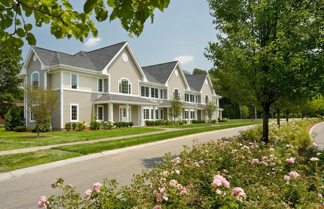 Row of beige townhome-style apartment buildings with landscaped lawns and pink flowers along a curved street.