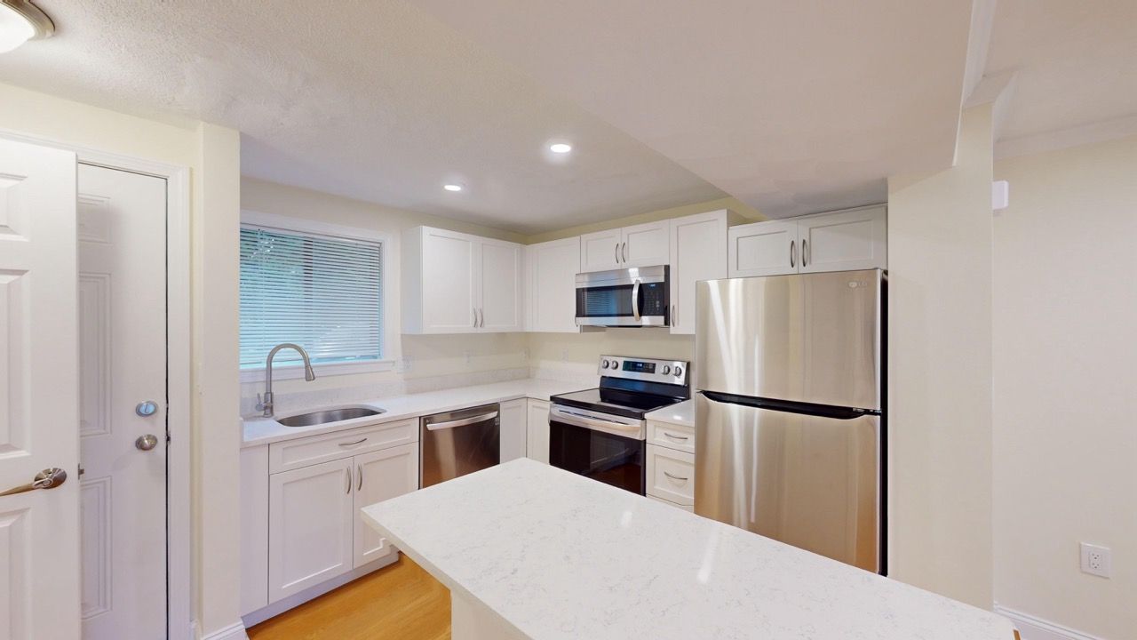 White kitchen with island, stainless steel refrigerator, oven, microwave, and sink under a window.