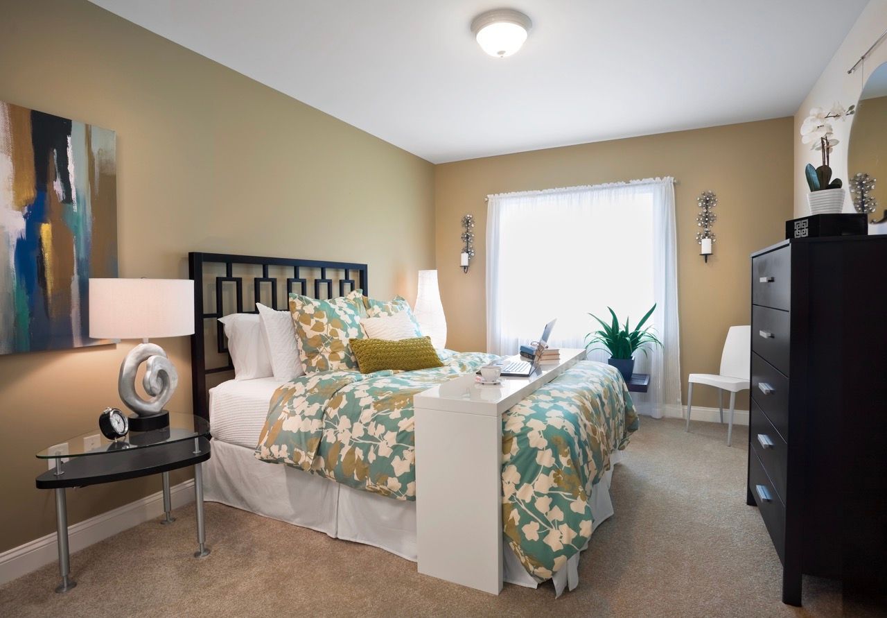 Bedroom in an apartment with a teal floral comforter, white furniture, and a window with sheer curtains.