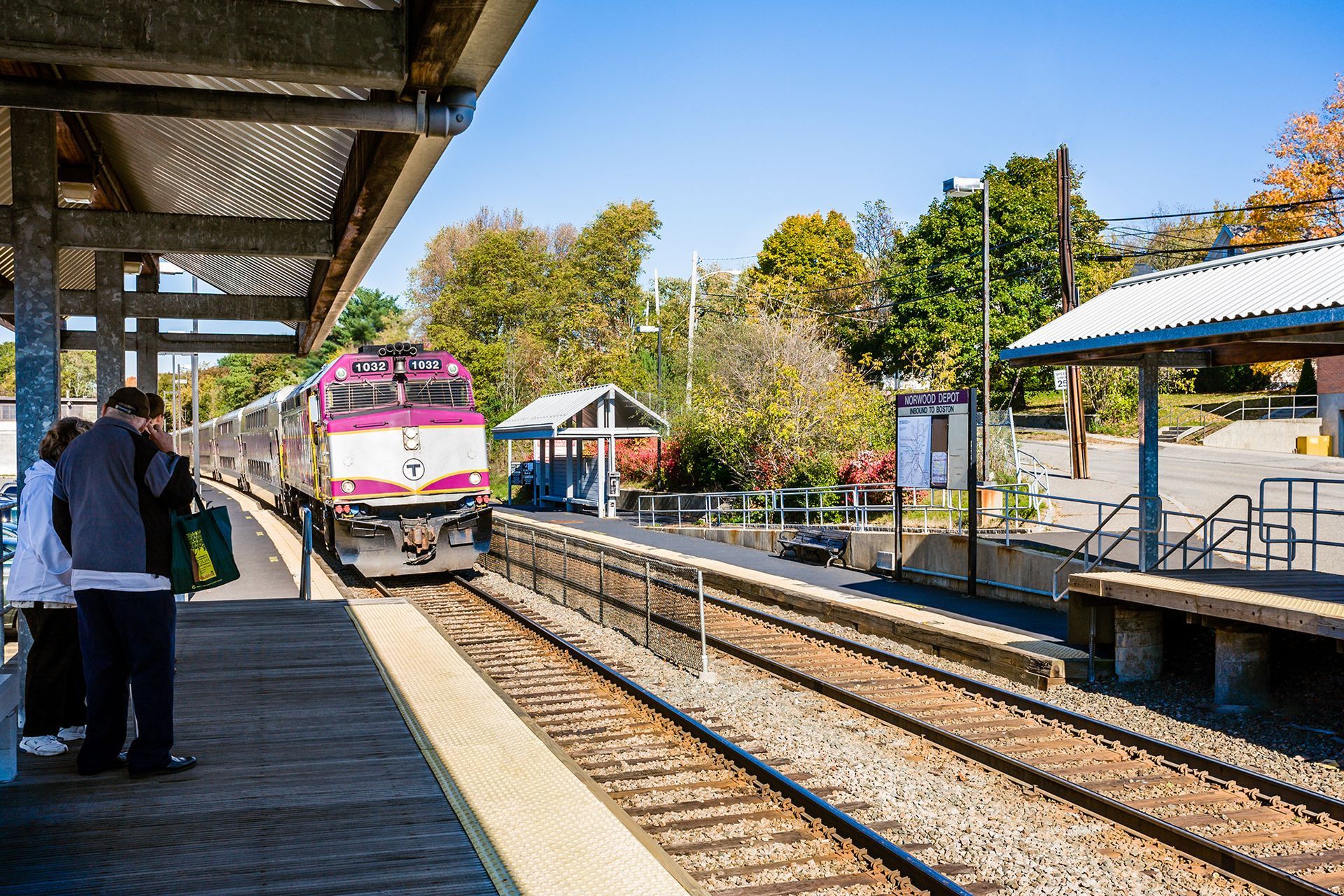 Pink-and-white train approaching a small outdoor station platform on a sunny day