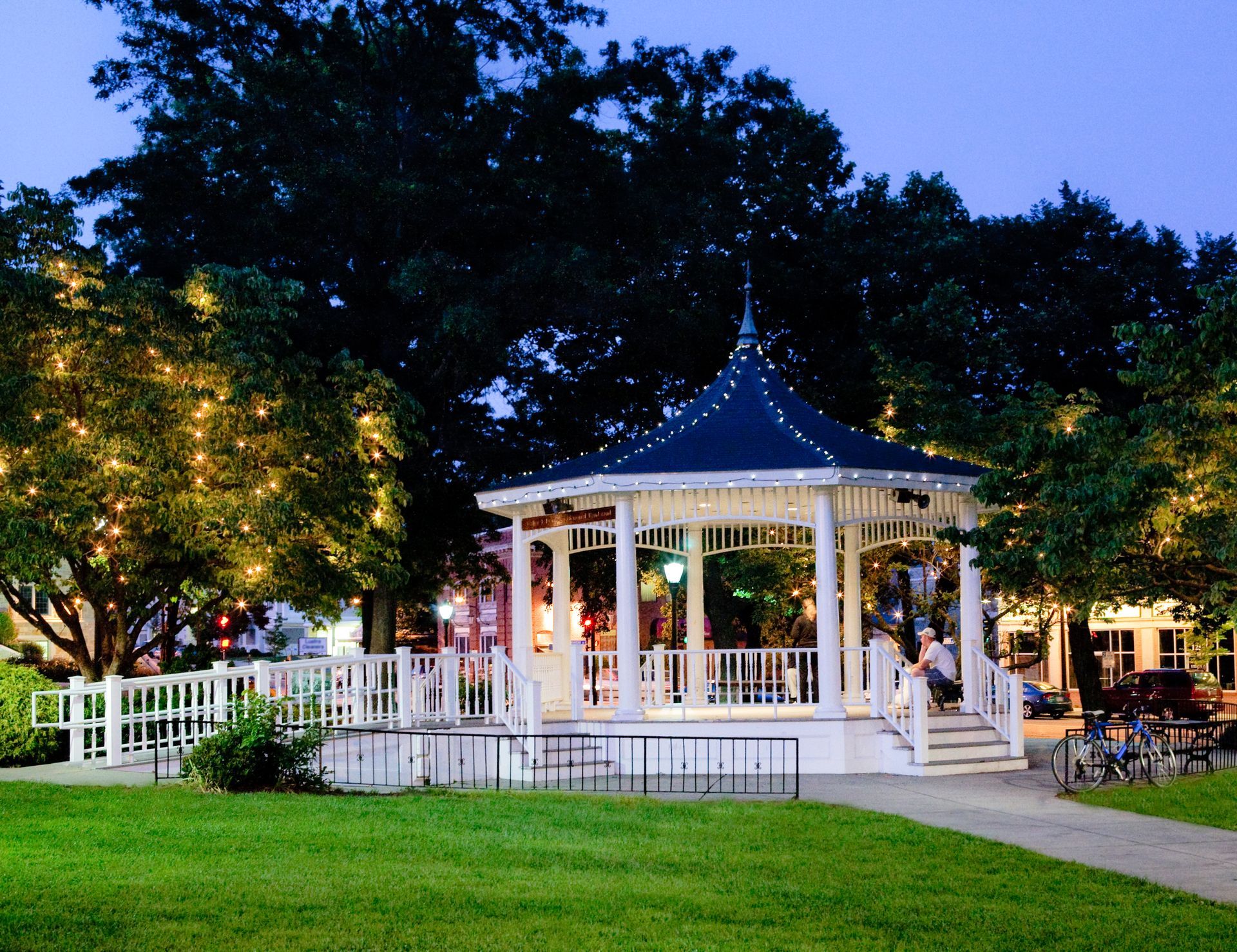 Lit white gazebo with blue roof in a park at dusk, surrounded by trees and green lawn