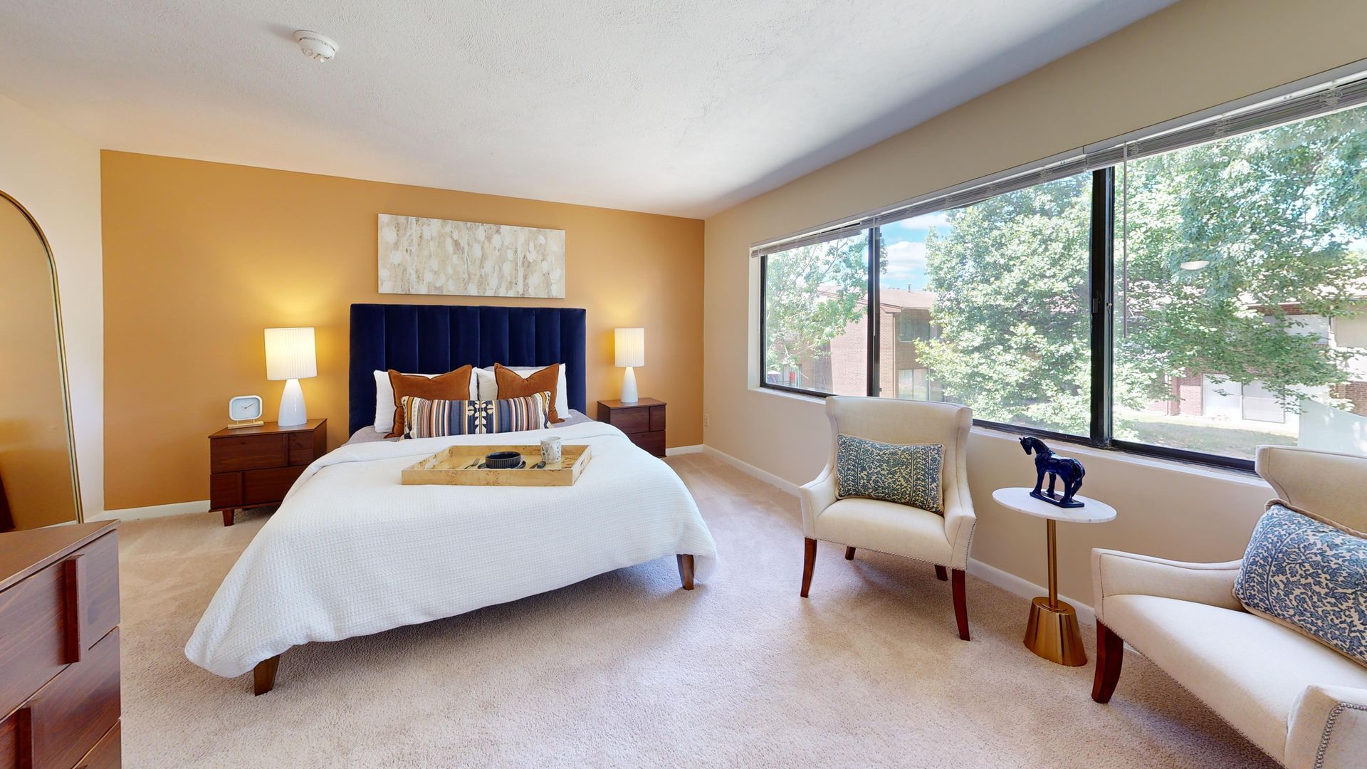 Bright bedroom with a bed, chairs, lamp, and large windows overlooking trees