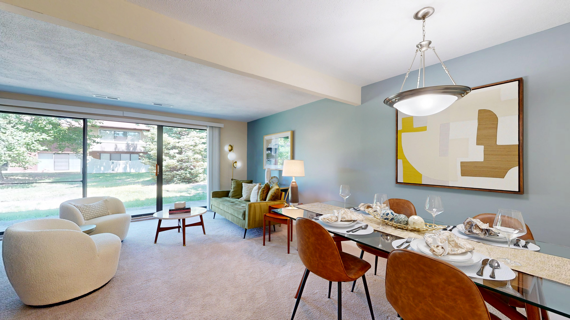 Bright modern dining and living room with large windows, white seating, and a set table beside a wall painting