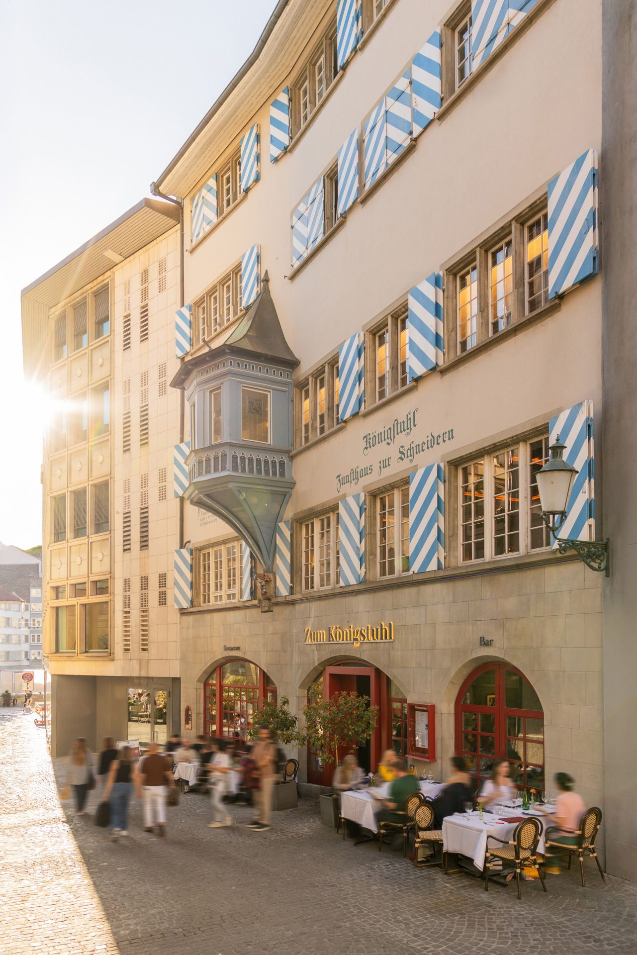 Sonnendurchflutetes Straßencafé neben einem hellbraunen Gebäude mit blauen Fensterläden und Bogenfenstern in einer europäischen Altstadt