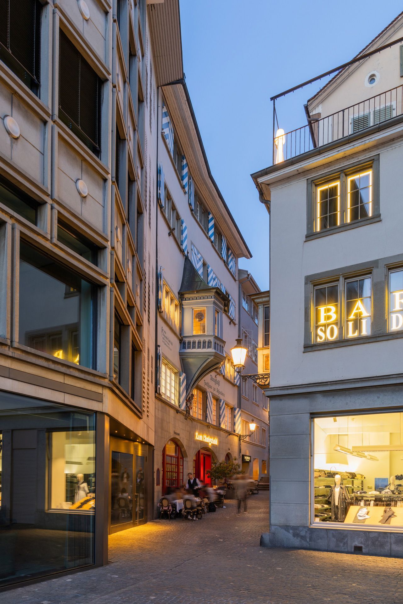 Enge europäische Straße in der Abenddämmerung mit beleuchteten Schaufenstern und historischen Gebäuden.