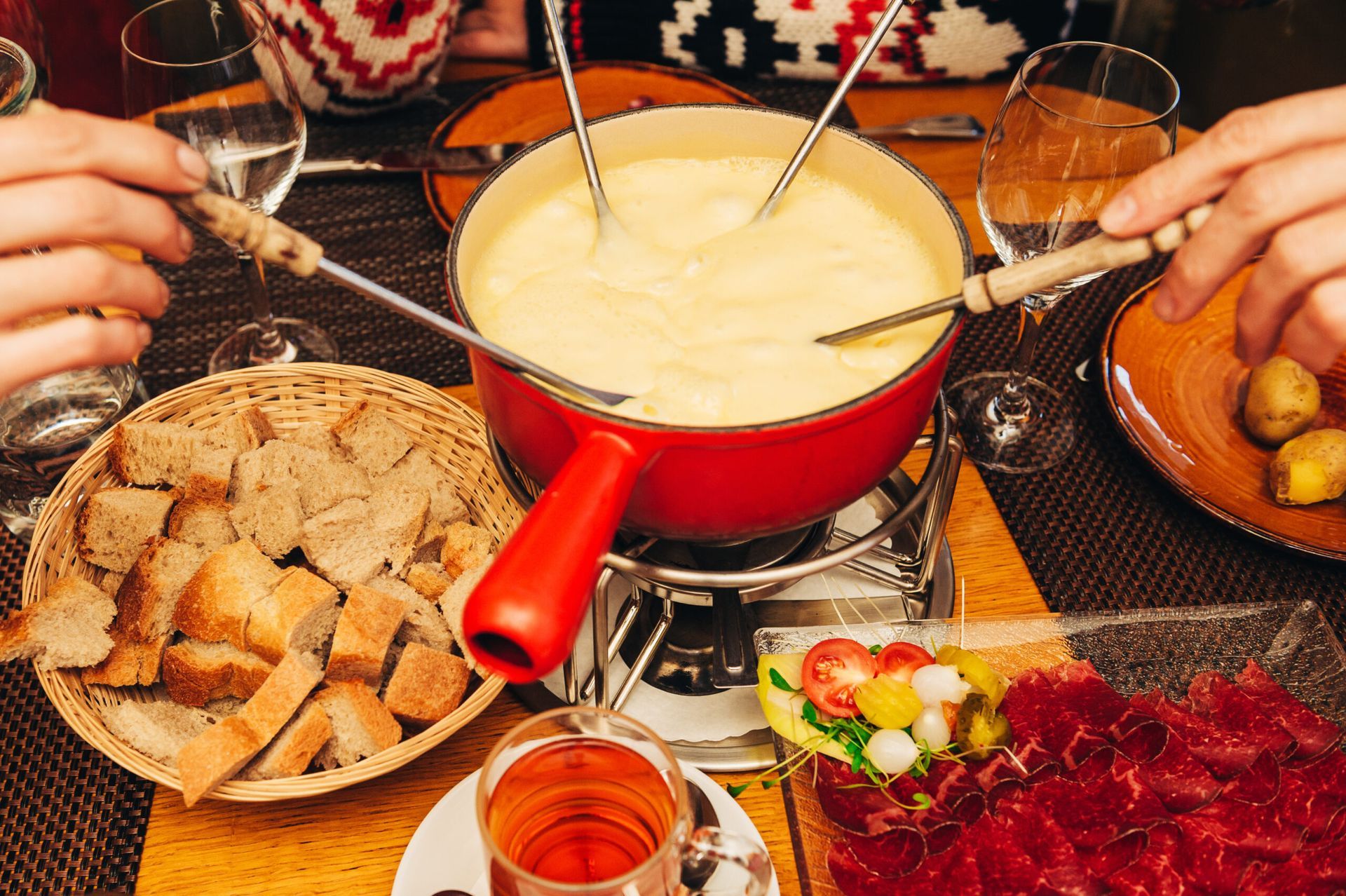 People dipping bread into a red fondue pot on a table with snacks and tea