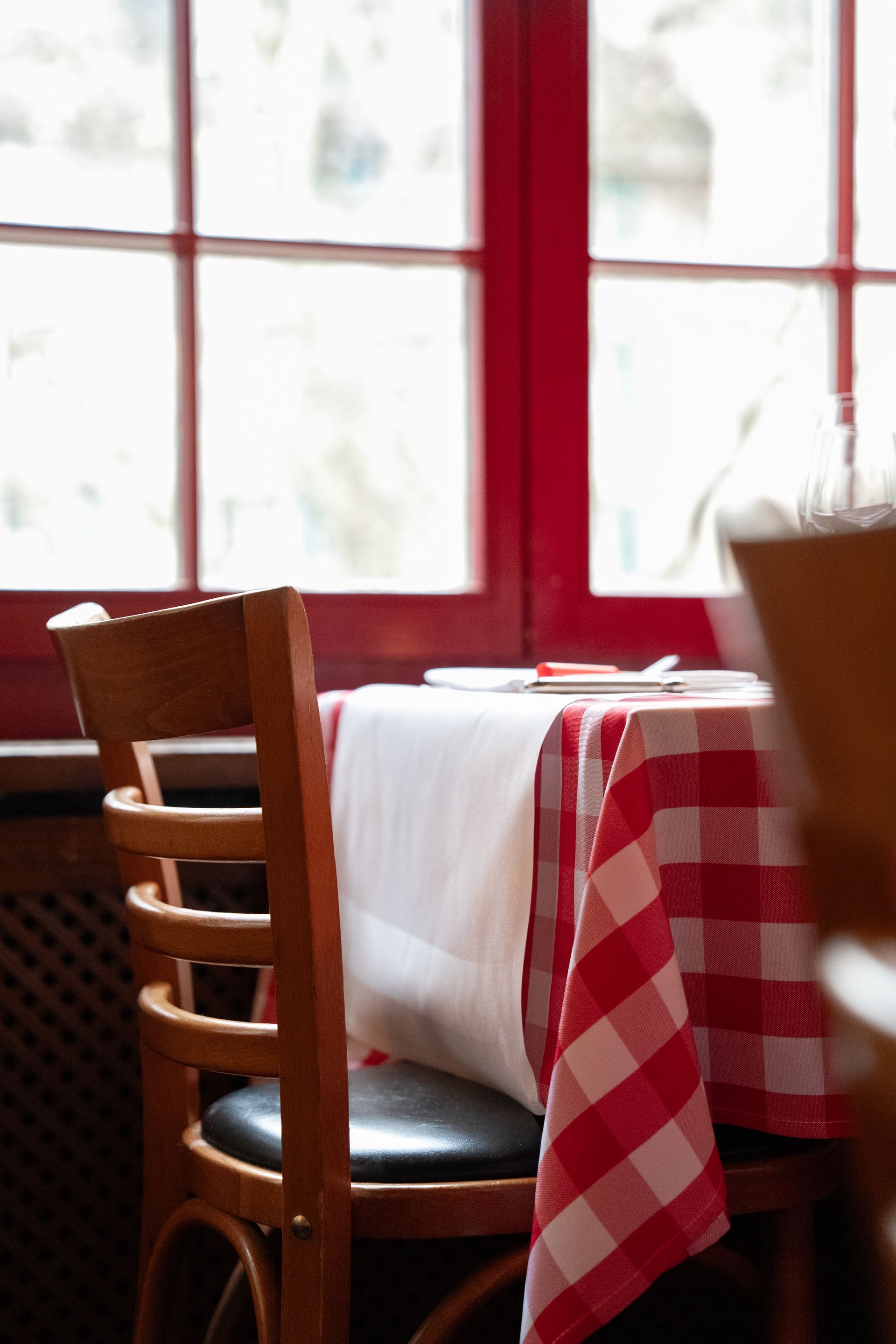 Gemütlicher Restauranttisch an einem rotgerahmten Fenster mit weißer Tischdecke und rot-kariertem Tischläufer.